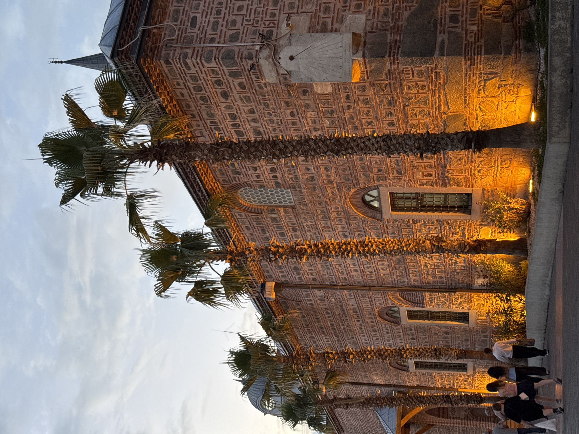 The image shows the exterior of the Dzhumaya Mosque in Plovdiv, Bulgaria, at dusk. The mosque is constructed of red brick and stone, with arched windows. Several palm trees line the front of the building, illuminated by spotlights. A sundial is mounted on the right side of the building. A group of people are walking away from the camera on a paved walkway in front of the mosque. The sky is cloudy and the overall lighting suggests it is late evening.