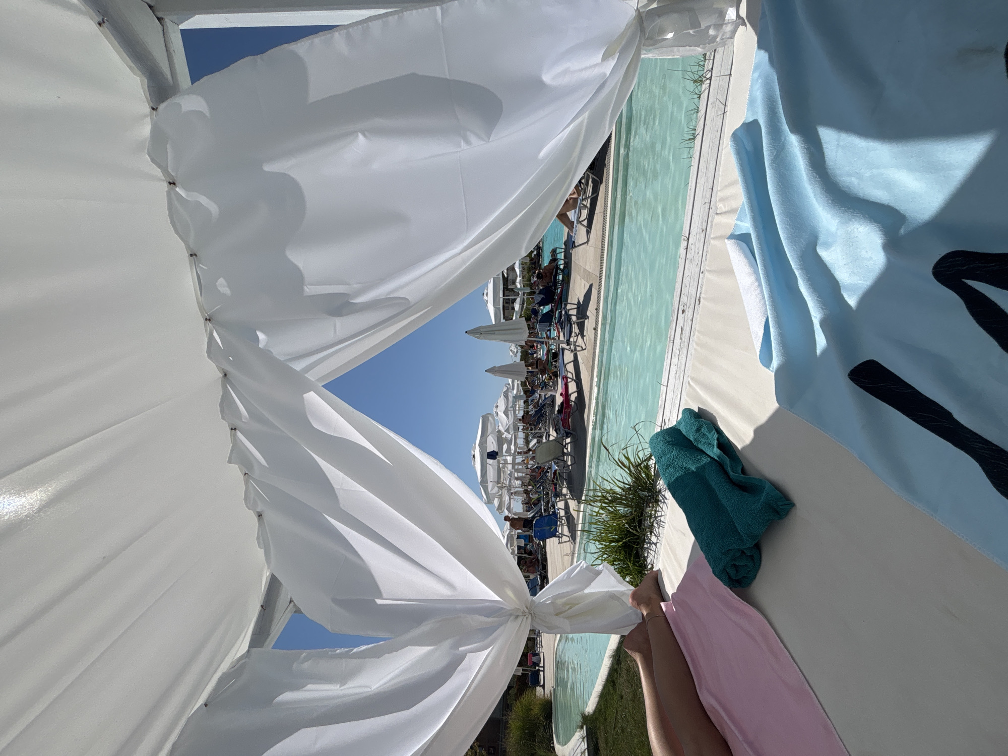 The image depicts a sunny day at a resort with a swimming pool in Byala, Bulgaria. In the foreground, a white canopy provides shade, with sheer white curtains draped to the side. On a lounge bed beneath the canopy, there are three towels: one pink, one teal, and one light blue with a black graphic design. The legs of a person are visible on the left side, lying on the pink towel. Beyond the bed, a bright blue swimming pool stretches across the scene, bordered by wooden decking and lush green grass. In the background, numerous beach umbrellas and lounge chairs are occupied by people, indicating a populated resort area. The sky is clear and blue.