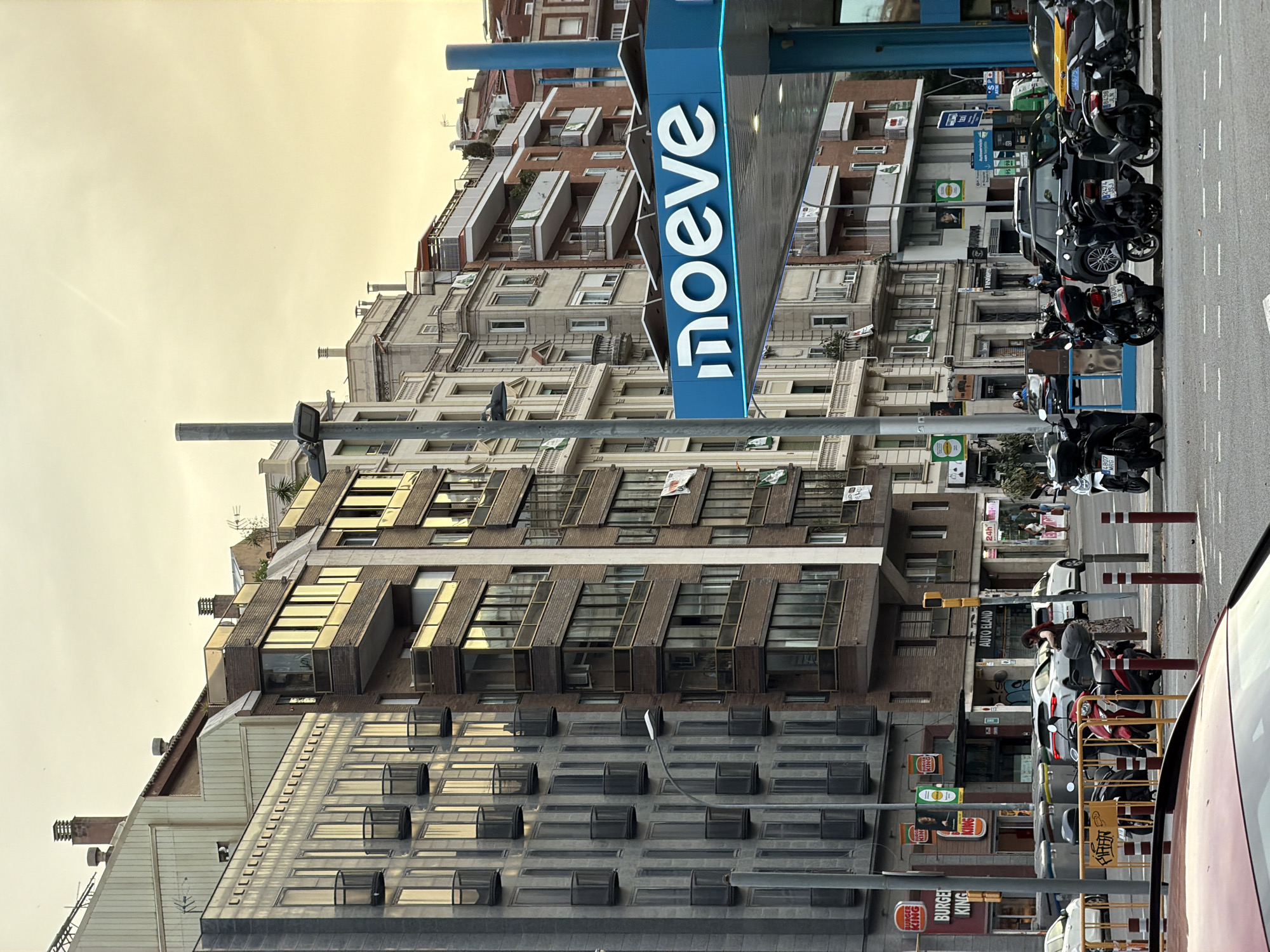 The image shows a street scene in Barcelona, Spain, with buildings lining both sides of the road and a prominent gas station canopy in the foreground. The gas station features a blue canopy with the white logo "moeve" in large lettering. Several motorcycles are parked on the sidewalk next to the gas station.
On the left side of the street, a Burger King restaurant is visible with its distinctive orange signage. Adjacent to it, another building has multiple floors with many windows and small balconies. Further down the street, the buildings are of varying architectural styles and heights, displaying a mix of modern and older construction. Some buildings have balconies with railings, and others feature large glass windows.
On the right side of the street, more multi-story buildings are visible, some with ornate architectural details and balconies. A yellow taxi is parked at the gas station. People are seen walking on the sidewalks and near the vehicles. The sky is overcast with a pale yellow hue, suggesting either early morning or late afternoon. The street is paved with asphalt, marked with white lines for traffic lanes and parking. The overall impression is of a bustling urban environment.