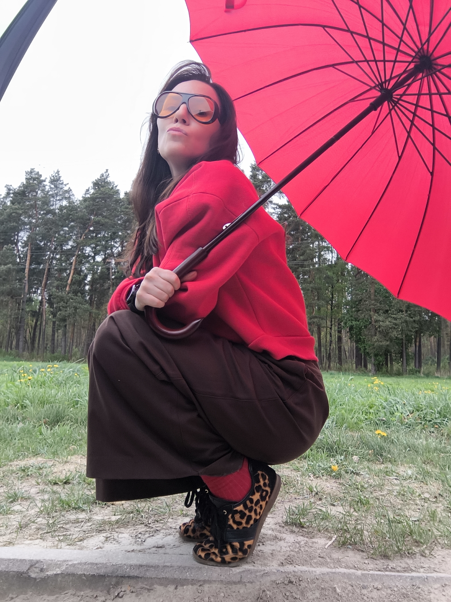 A young woman is crouching on what appears to be a concrete barrier, likely near a park or wooded area. She is wearing a red sweatshirt, brown pants, red socks, and leopard print sneakers. She is holding a red umbrella. She has long brown hair with a streak of grey near her forehead and is wearing oversized sunglasses. In the background, there are trees and a grassy area with yellow flowers. The setting is likely a park area within the city of Riga, Latvia. The picture is taken from a low angle, looking slightly up at the subject. The sky is overcast.