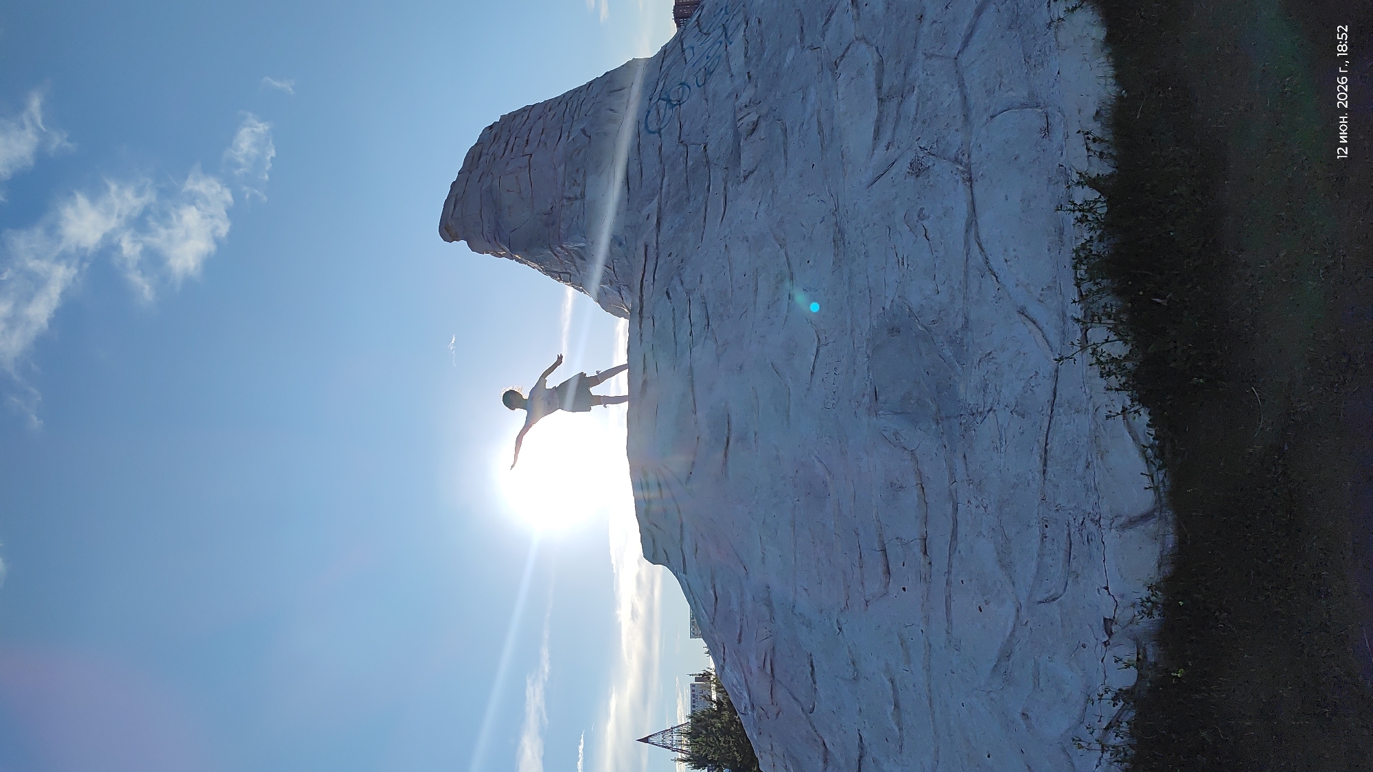 A child, silhouetted against the bright sun, stands with arms outstretched on top of a large, textured, light-colored rock formation. The child appears to be wearing a skirt and socks. The sun is positioned in the upper left quadrant of the image, casting rays and creating lens flare. The rock formation dominates the foreground and midground, with a smaller peak to the right. In the background, a clear blue sky with scattered white clouds is visible. To the left, a modern-looking spire-shaped structure and some trees can be seen, suggesting an urban environment. The bottom of the image shows dark grass and vegetation. A timestamp at the bottom right reads "12 июня. 2026 г., 18:52". This scene is located in the city of Astana, Kazakhstan.