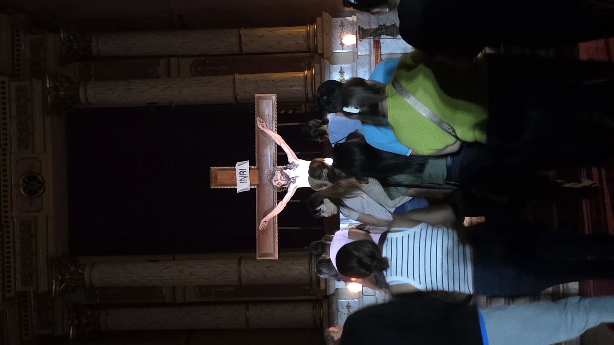 A photograph shows a vertical image of a crucifix placed within a religious setting. The crucifix features the figure of Jesus Christ, and above his head, there is a sign with the inscription "INRI".
The scene is set in a room with notable architectural details including pillars with capitals. People are gathered around the crucifix; their presence suggests a religious ceremony or observation. The room is illuminated, but the backdrop behind the crucifix is dark, which suggests it is in a church.
The city and exact location are not discernible from the photograph.