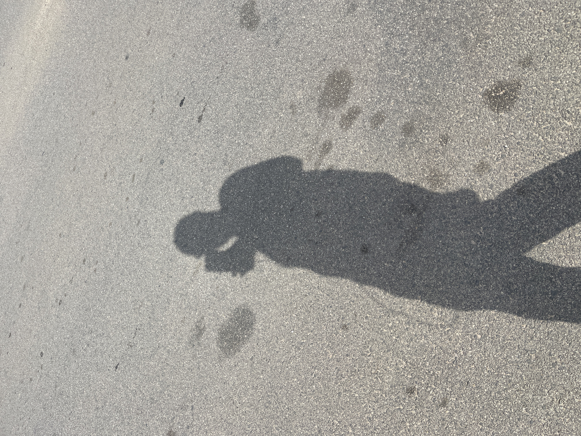 The image shows the shadow of a person standing on a paved surface. The shadow suggests the person is holding something near their face. The location appears to be an outdoor area with an asphalt or concrete surface. No identifying details place the scene in Gosa, Nigeria.