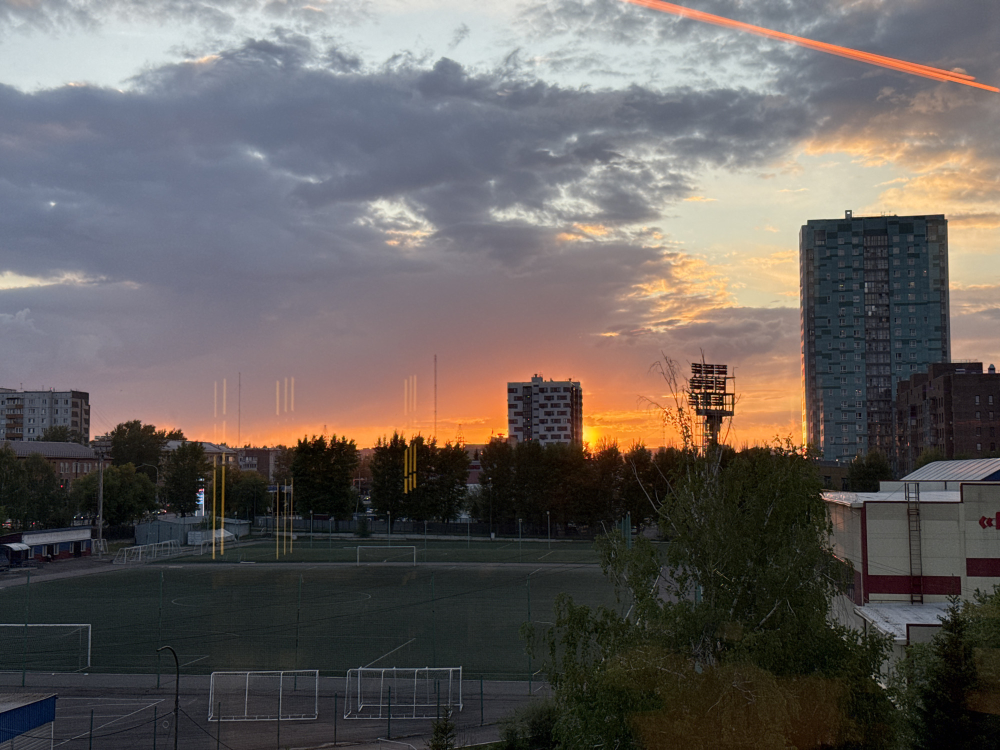 The image depicts a cityscape at sunset. In the foreground is a soccer field with multiple goalposts. Behind the field, there's a row of trees, followed by various buildings. Two multi-story buildings stand out against the sunset sky. The sky is a mix of dark, cloudy areas and orange hues where the sun is setting. A reddish line is visible in the upper right portion of the sky, likely from an airplane contrail. The location is Krasnoyarsk, Russia. There are no people visible in the image.