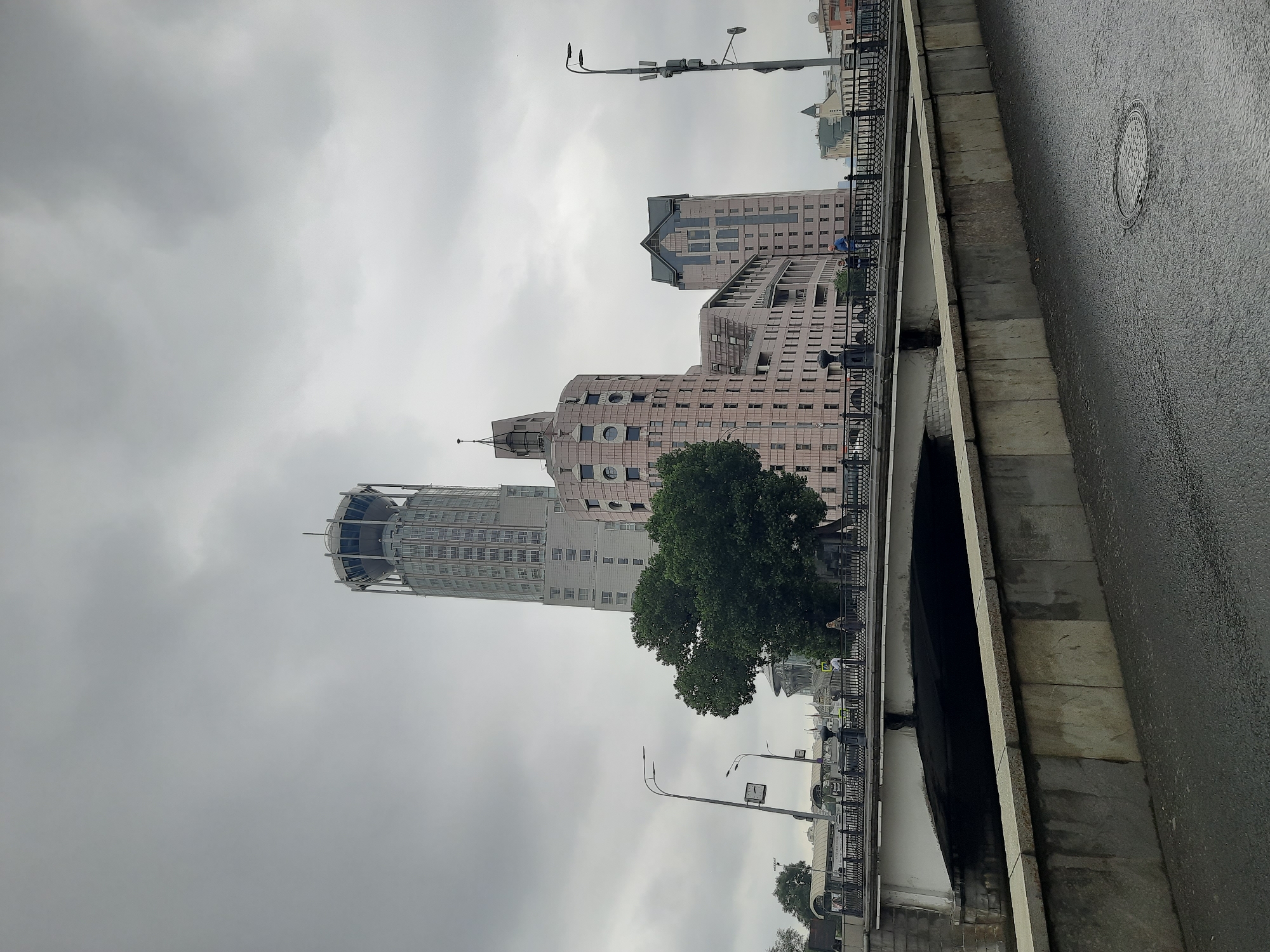 A wide-angle shot captures a cloudy, overcast day in an urban setting, possibly Moscow. In the background, several modern buildings rise prominently. The tallest, a slender, pale skyscraper with a distinctive circular observation deck at its summit, dominates the left side of the frame. To its right, a cluster of pinkish-hued buildings with rounded corners and numerous windows creates a unique architectural ensemble. Further to the right, another taller building with a pointed roof and a dark gray facade stands. In the mid-ground, a bridge spans a body of water, its white underside contrasting with the dark water below. People are visible walking on the bridge. A large, lush green tree is positioned between the bridge and the pink buildings. The foreground shows a wet, asphalted surface, suggesting recent rain, with a manhole cover visible on the right. Streetlights and lampposts are present along the bridge and the street. The sky is filled with heavy, gray clouds, creating a somber atmosphere.