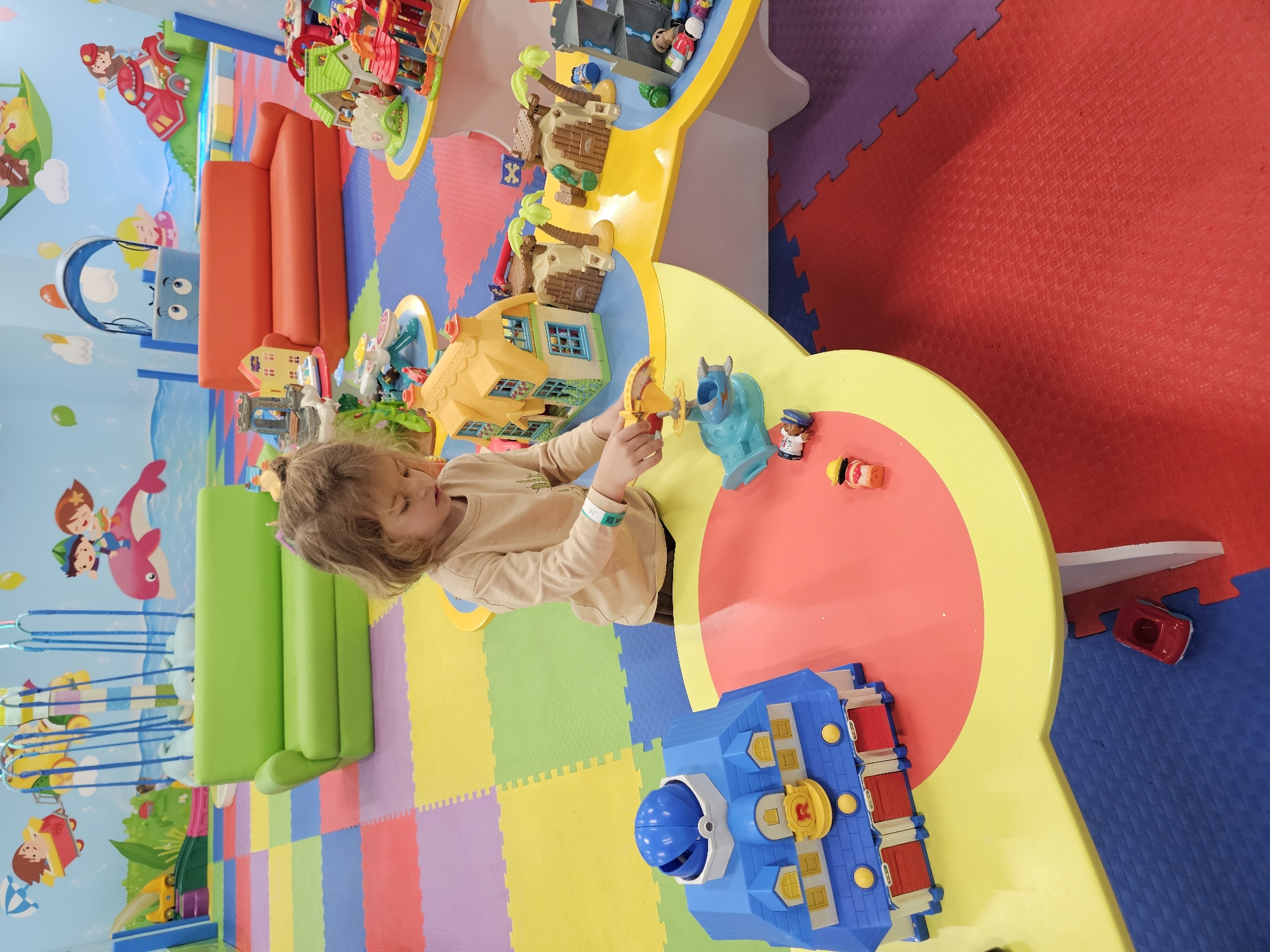 A child with blonde hair is playing with toys on a yellow and red table in an indoor play area. The room has colorful interlocking floor mats and a mural on the wall. The location is Khimki, Russia.