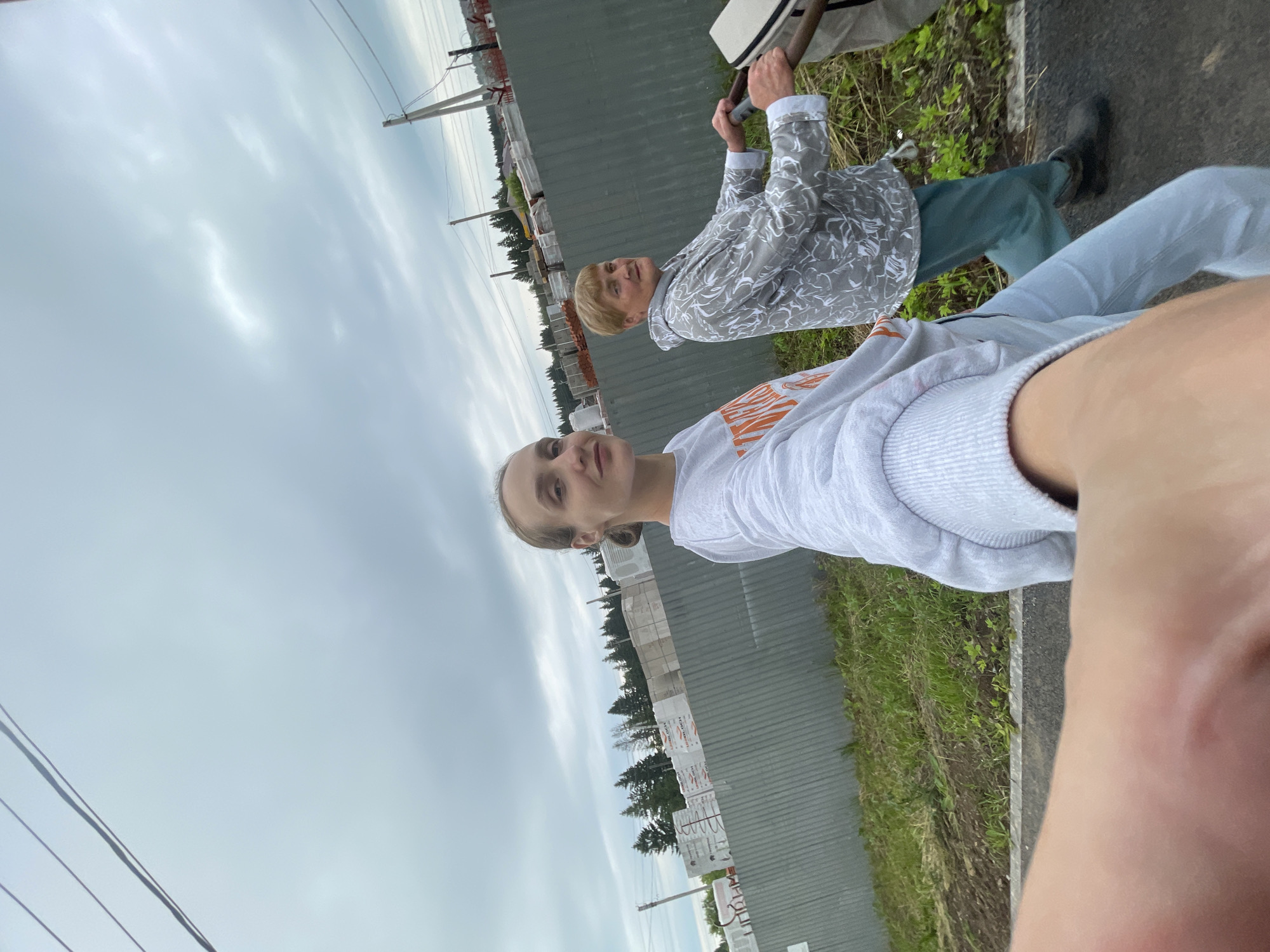 In this image, two people are outdoors with a cloudy sky overhead. In the foreground, a person is partially visible, appearing to be holding a phone to take a picture, with their arm and shoulder in frame. They are wearing a light gray sweatshirt with orange writing that is partially obscured.
Behind them, an older person is visible, looking towards the camera. They have short, blonde hair and are wearing a patterned grey and white jacket over a white shirt, and teal-colored pants. Their hands are clasped around a long, thin object that appears to be part of a cane or support.
The background shows a gray, corrugated metal structure that might be a fence or part of a building. To the right of this structure, there is a strip of green grass and some vegetation. Beyond that, a road or pathway is visible. In the distance, there are trees and what appear to be construction materials stacked in wrappers with some visible text that cannot be clearly deciphered. Electrical wires are also visible in the upper portion of the image, stretching across the sky. The overall impression is of a casual, outdoor setting, possibly near a construction site or an industrial area. The lighting suggests it is daytime, but the cloudy sky indicates overcast weather.
