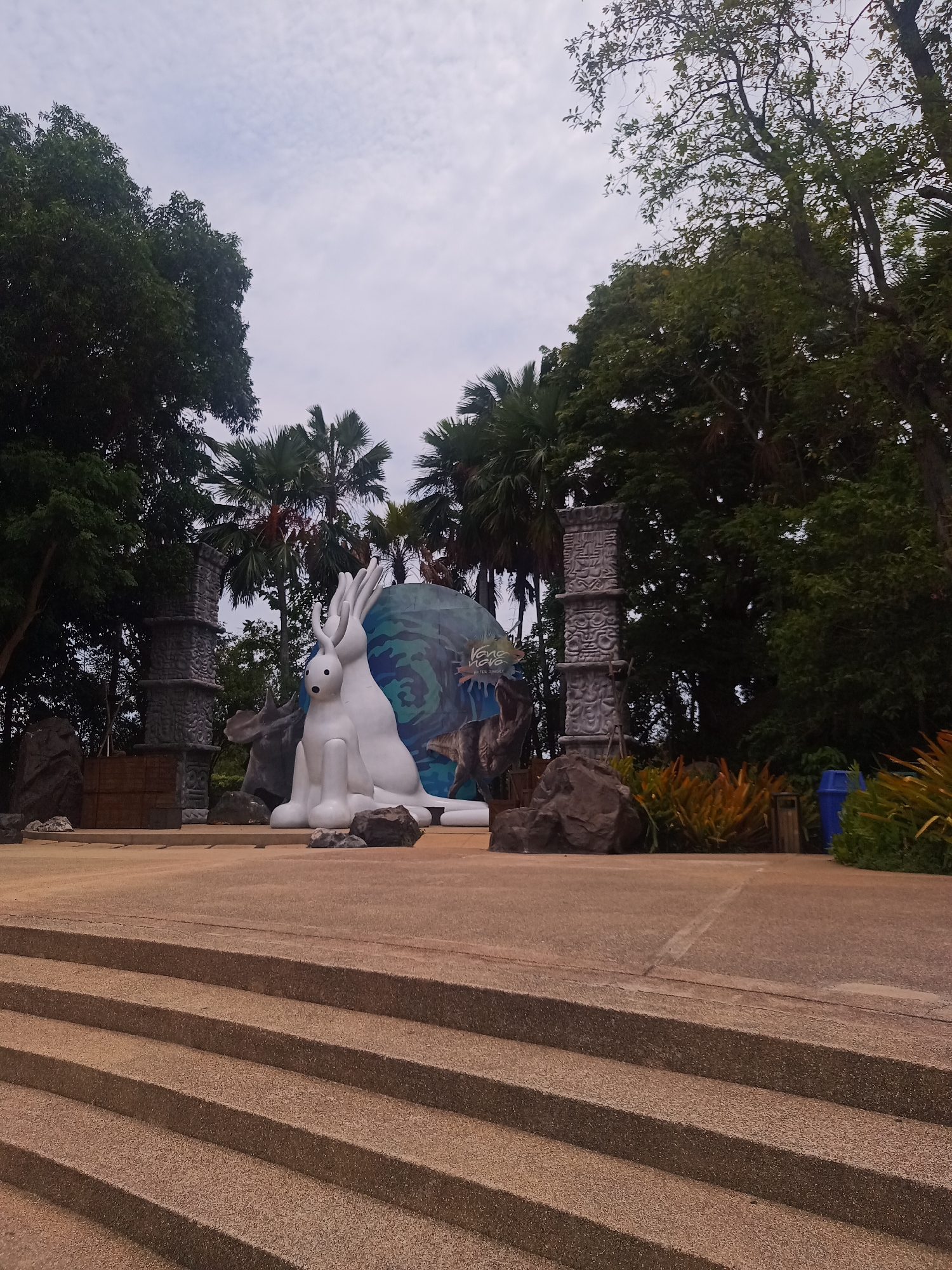 The image shows a tourist attraction in Hua Hin City Municipality, Thailand. In the background are palm trees, trees, and a cloudy sky. In the middle ground, there are decorative stone columns with carved designs and a large white sculpture of two rabbits positioned in front of a backdrop with a blue and green design, possibly representing a globe or water theme, with a sign that reads Vana Nava water Jungle. In the foreground, there are stone steps leading up to the platform where the sculpture is located. There are also rocks and plants on the platform. A blue bin and a trash can can be seen behind one of the stone pillars on the right side. The scene is set in what appears to be an outdoor park or attraction.