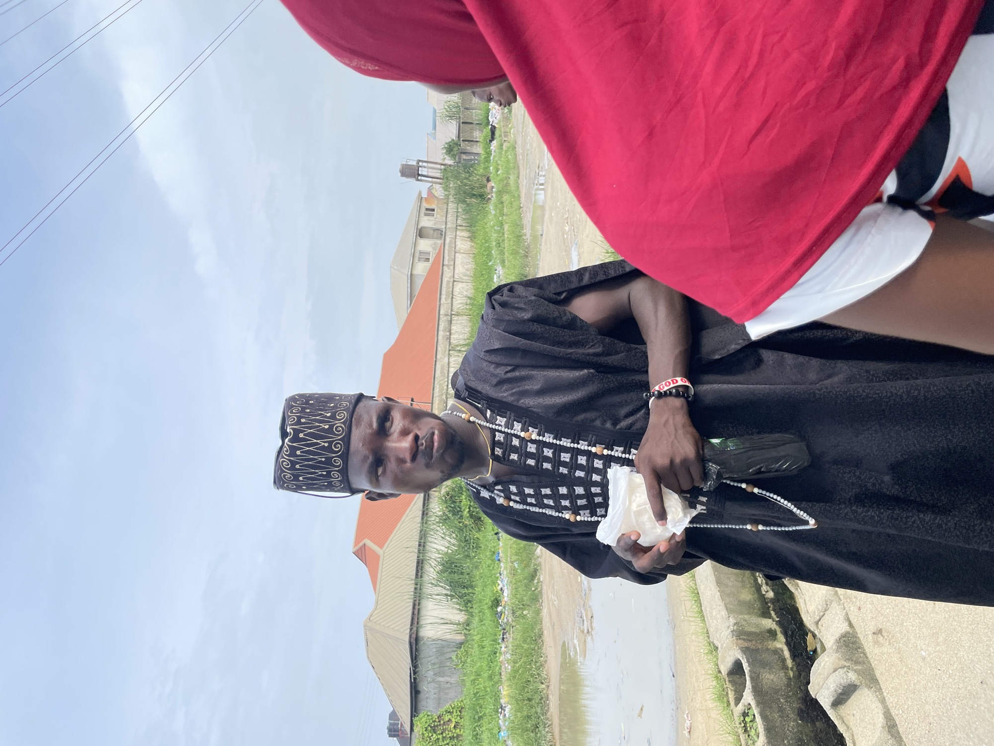 A man wearing a traditional black agbada and a patterned cap stands on a street in Ado, Nigeria. He is holding a white object in his hands. The environment shows urban buildings, green vegetation, and stagnant water on the side of the road. Part of another person's red shirt is visible in the foreground. Overhead electric lines are also present.