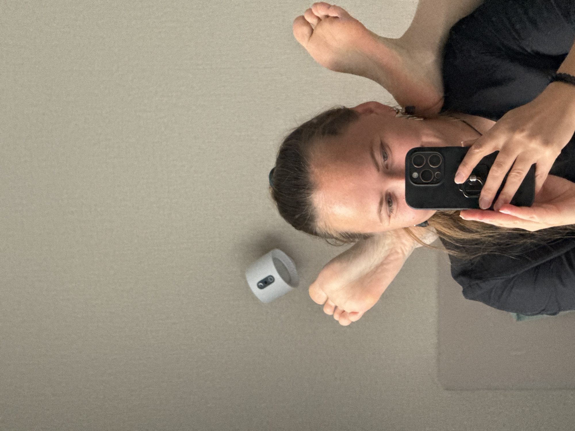 A woman is holding a smartphone taking a picture of herself. The woman has brown hair pulled back, and is wearing a black shirt. Her feet are in the upper left corner of the photo. There is a white object with two black dots on it near her feet. The background is a grey wall. There is no evidence to suggest the image was taken in Izhevsk, Russia.