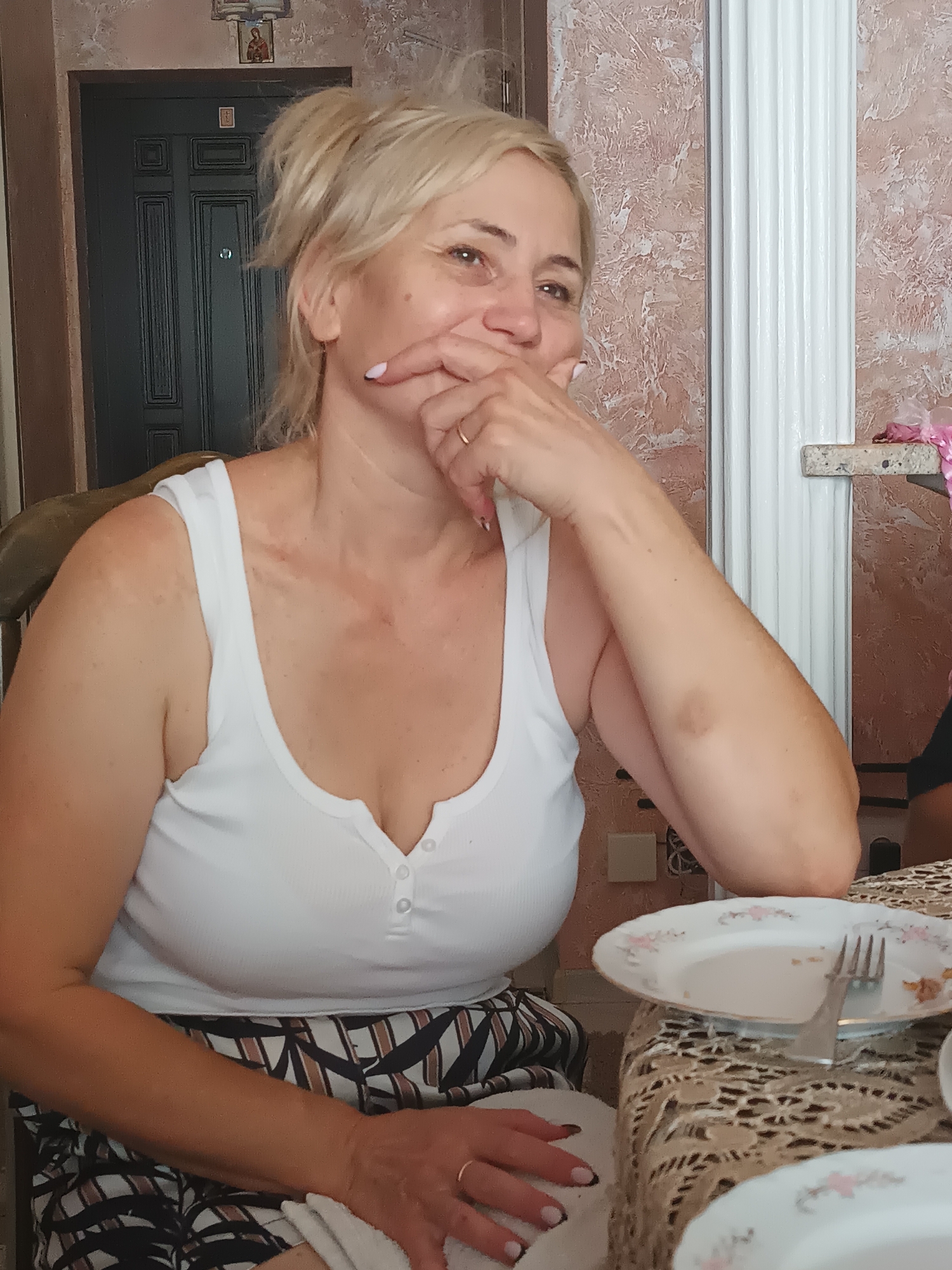 The photograph captures an indoor scene, likely a dining area, with a woman in the foreground. She has blonde hair pulled back and is wearing a white tank top. Her right hand is near her face, with her fingers touching her mouth. She is seated at a table covered with a patterned tablecloth. On the table, there are plates and cutlery, suggesting a meal has recently taken place or is in progress. The background shows a door with a decorative element above it and textured walls. The overall setting appears to be a domestic environment. The location is identified as Durlești, Moldova.