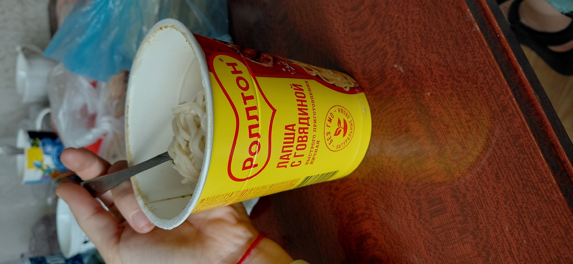 The image shows a person holding a spoon in a cup of instant noodles. The cup is yellow with red lettering that reads "РОЛЛТОН" (Rollton) and "ЛАПША С ГОВЯДИНОЙ" (Noodles with Beef). There is also a "БЕЗ ГМО" (GMO-free) and "НАТУРАЛЬНЫЕ ОВОЩИ" (Natural Vegetables) symbol on the cup. The noodles are light brown and appear to be cooked. The cup is sitting on a wooden table. In the background, there are several mugs and plastic bags. This image was taken in Svetlogorsk, Russia.