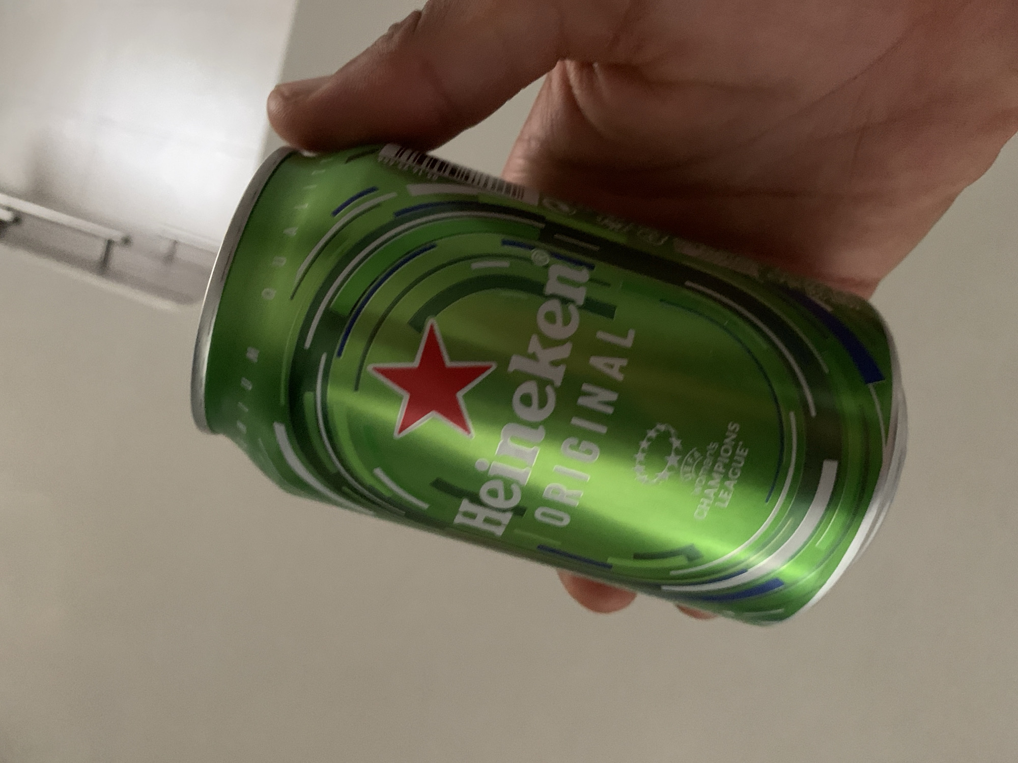 A Heineken Original beer can is held in a person's hand. The can is green with a red star and white text. Below the Heineken logo, "ORIGINAL" is visible, and further down, a UEFA Women's Champions League logo is present. The background is a plain, light-colored wall. The scene is indoors, and there is no indication of it being in Barcelona, Spain.