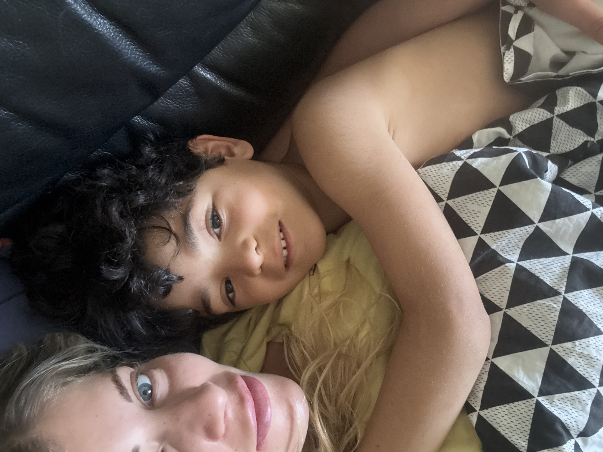 The image shows two people lying down together. One person, a child with dark curly hair, is lying on a yellow shirt. They are looking at the camera and smiling slightly. The other person, an adult, is lying beneath the child. The adult has blonde hair and is partially visible. The scene is set indoors, with a dark leather surface visible in the background, and a black and white triangular patterned blanket. Location is not possible to verify as Rousset, France.
