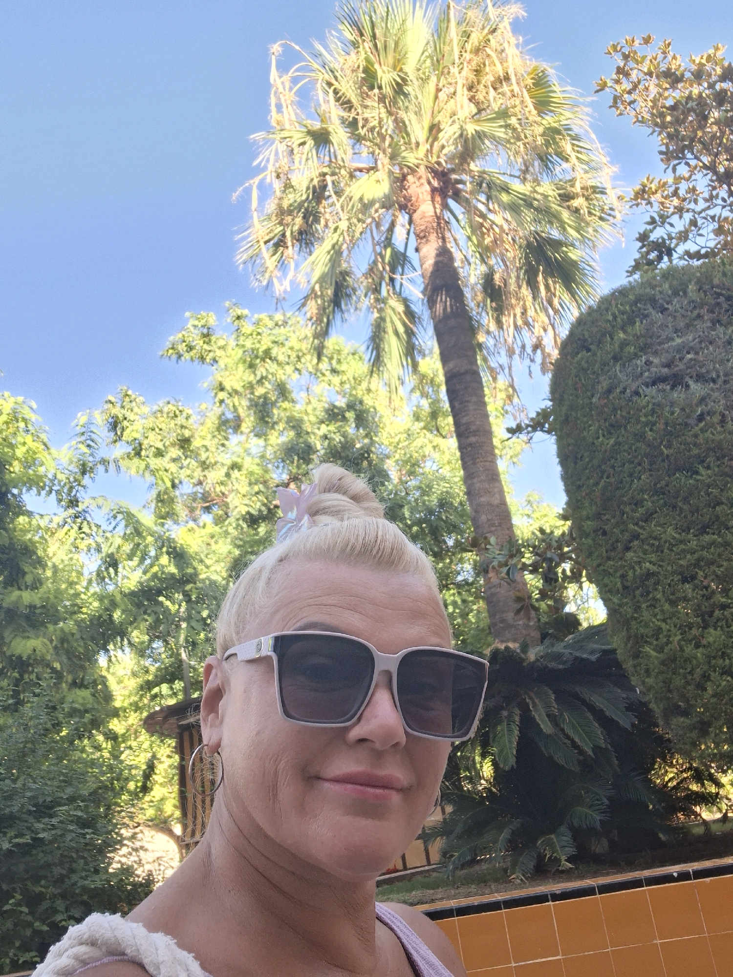 A woman with blonde hair styled in a bun and a pink butterfly clip is taking a selfie outdoors in a garden with palm trees and lush green foliage under a clear blue sky. She is wearing large, square-framed sunglasses with white accents and a hoop earring in her left ear. The woman is also wearing a light-colored top with a thick rope strap. The background features a tall palm tree, other trees, and a neatly trimmed hedge. A portion of an orange tiled surface is visible in the lower right corner. The scene suggests a sunny day, possibly in a Mediterranean climate, consistent with the city of Torrevieja, Spain, which is known for its coastal gardens and palm trees.