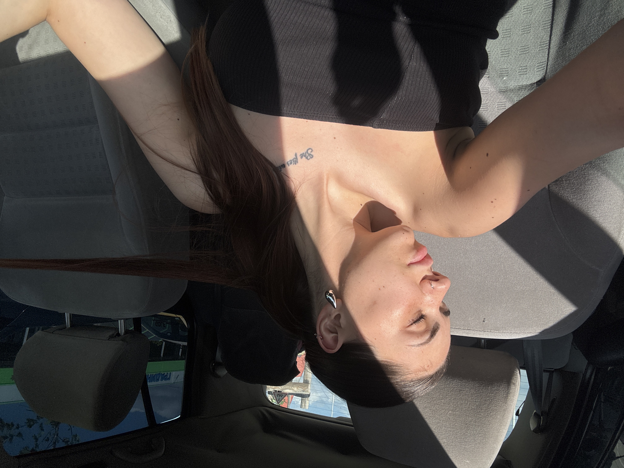 A young woman is depicted in a medium shot, seated in the front passenger seat of a car, facing slightly towards the viewer's left. Her eyes are closed or mostly closed, and her head is tilted. She has dark hair styled in a ponytail, with a long strand extending over the back of the seat to the right. Her skin shows freckles and moles. She wears a nose ring on her right nostril and two silver-colored earrings in her left ear: a teardrop-shaped stud and a small hoop. On her left collarbone, a tattoo partially reads "She flies with". She is wearing a black, ribbed strapless top. Strong sunlight enters from the right, casting sharp shadows across her face, chest, arm, and the grey fabric car seats. Through the car's rear window, a building with green Cyrillic text on a white background is visible, clearly showing "ГРАДИН" (GRADIN). Blue sky with some clouds and faint tree foliage are also visible in the background. The presence of the Cyrillic text "ГРАДИН" on a commercial building identifies the location as Sofia, Bulgaria.
