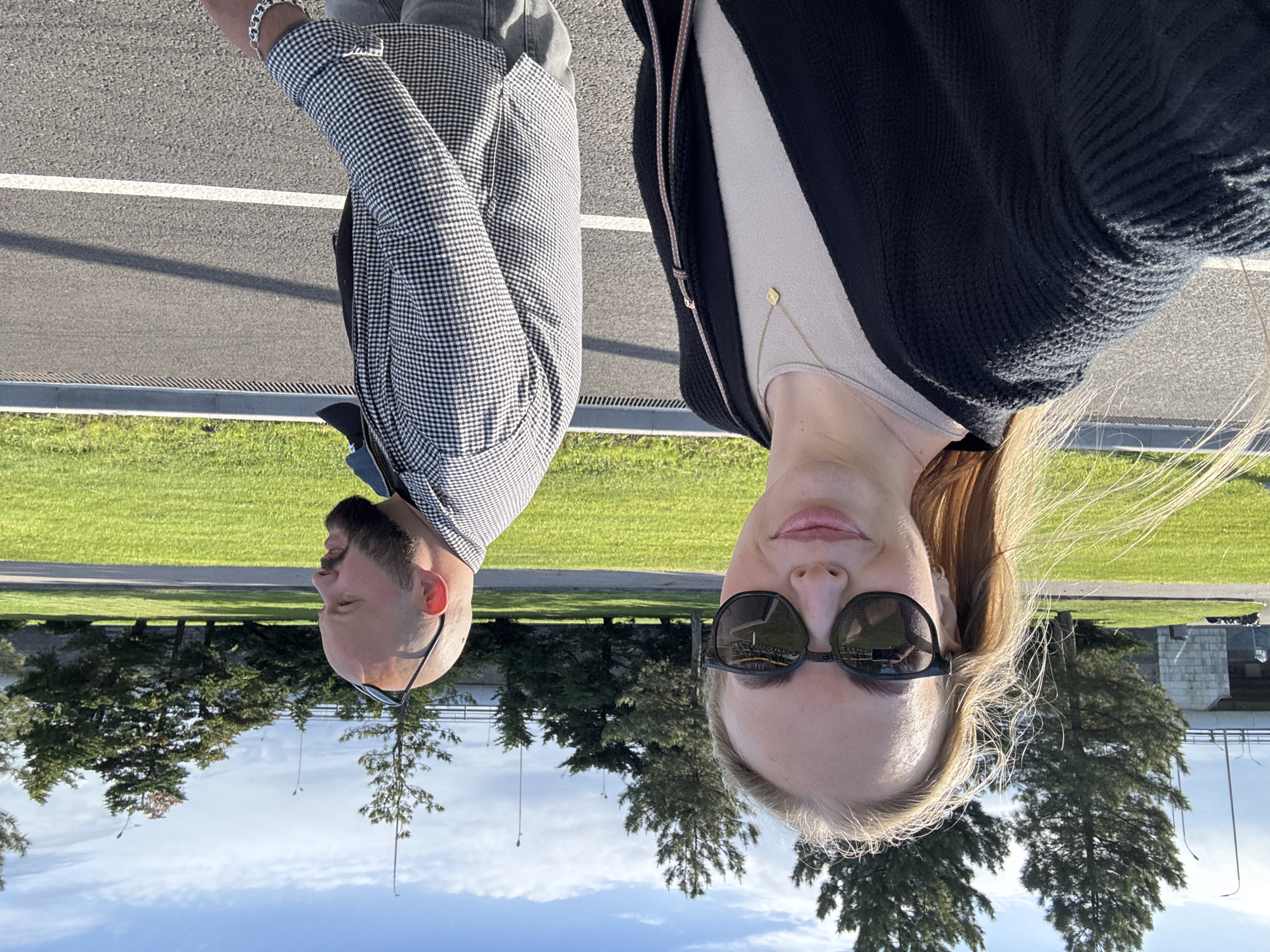 Two people are in the image: a man and a woman. The man is on the left, wearing a checkered jacket and glasses. He has a beard and is bald. The woman is on the right, wearing sunglasses, a light-colored top, and a black cardigan. She has blonde hair. Both are standing on a paved surface with a white line. There is grass on either side of the pavement, and trees are visible in the background. The image is taken outdoors, and the sky is visible at the bottom. The location is possibly in or near Sirius, Russia. The image has been rotated 180 degrees.