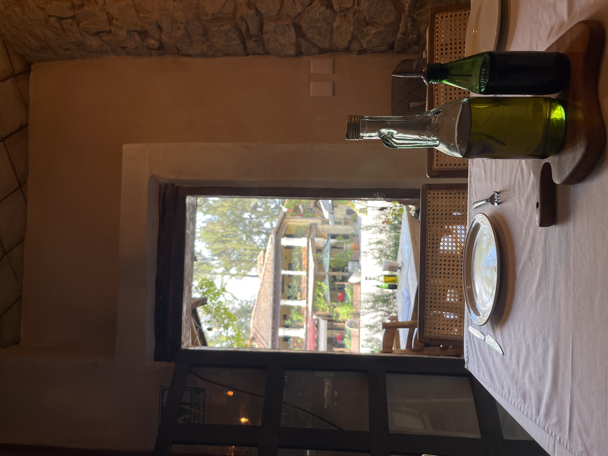 The image captures a dining scene in what appears to be a restaurant or similar establishment, situated in the city of Domingo Arenas, Mexico. The view through the window reveals a multi-story building with architectural features typical of Mexican design, complete with balconies, terracotta roofs, and visible greenery. Inside, the focus is on a table setting adorned with a light-colored tablecloth. Place settings include plates and cutlery. Prominently featured are two glass bottles containing liquids, placed on a wooden serving board. The lighting is soft and diffused. The overall ambiance suggests a quiet, possibly daytime, setting. The physical environment includes stone walls and furniture with woven elements. No people are visible in the shot.