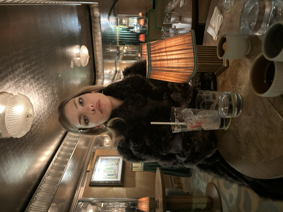 A woman with blonde hair and a dark furry coat sits at a table in a dimly lit bar. A lampshade emits a warm glow. Drinks are scattered across the table, including a half-empty glass with a straw. Behind the woman is a wall with ornate molding and sconce lighting. A framed picture hangs on the wall. The scene evokes a sense of intimacy and relaxation.
