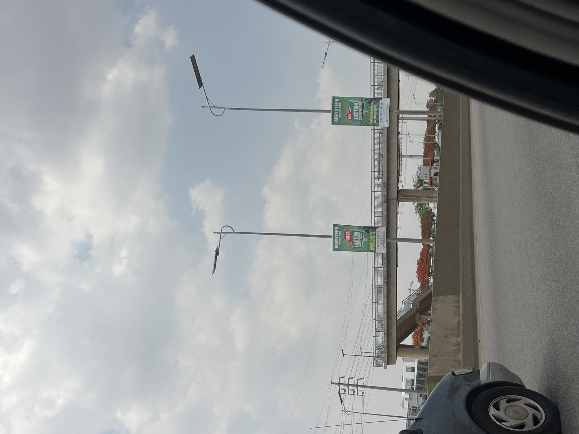 This media file captures a roadside scene in Kuchingoro, Nigeria, during what appears to be an overcast or partly cloudy day. The image is taken from a low angle, possibly from inside a vehicle, looking towards an elevated pedestrian bridge or overpass.
Two tall streetlights, each equipped with what looks like a solar panel, extend upwards from the ground. Attached to poles near the bridge are two large green advertising banners. The text on these banners, as confirmed by OCR, reads "ABEYSTEPH PRINTING PRESS" (or "PROITINS PRESS") and encourages potential customers to "BUY ELECTION CAMPAIGN AND BRAND IT AT NO COST." Below this, "WUSE OFFICE" and a phone number "09135338533" are prominently displayed.
Beneath the elevated structure, various power lines are visible, connecting to utility poles. In the background, beyond the bridge, some buildings and trees with striking reddish-orange flowers can be seen. The asphalt road surface is visible, and the front right wheel and part of the bumper of a car are visible in the lower right corner, suggesting the viewer is in or near a vehicle. The sky is predominantly gray with scattered white clouds. The scene depicts a functional urban or semi-urban environment with typical infrastructure.