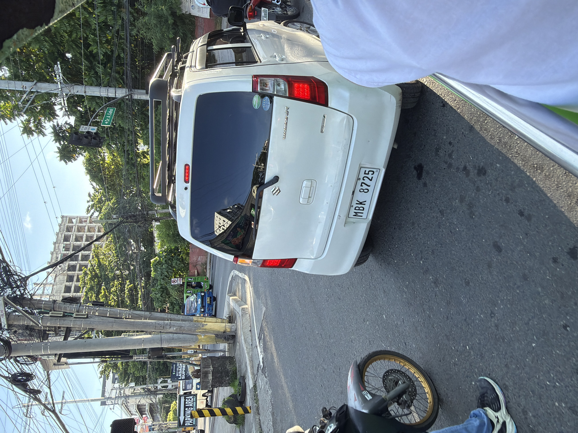 This image captures a street scene in Davao City, Philippines. A white Suzuki Wagon R is in the foreground, facing away from the camera, with its license plate clearly visible as MBK 8725. A motorcycle is to the left of the car, and the lower half of a person wearing blue jeans and black shoes is visible, suggesting they are on the motorcycle. In the background, a construction site with a partially built, multi-story building is visible amidst lush green trees and a clear blue sky. A traffic light hangs over the intersection, and a street sign indicates "Dr. A. Gahol." Several utility poles with a dense network of wires are prominent features of the urban landscape. A blue and green jeepney or similar public transport vehicle is also visible in the middle ground, adding to the bustling city atmosphere. The overall impression is a snapshot of daily life in a busy Philippine city, with a mix of modern infrastructure, ongoing development, and common modes of transportation.