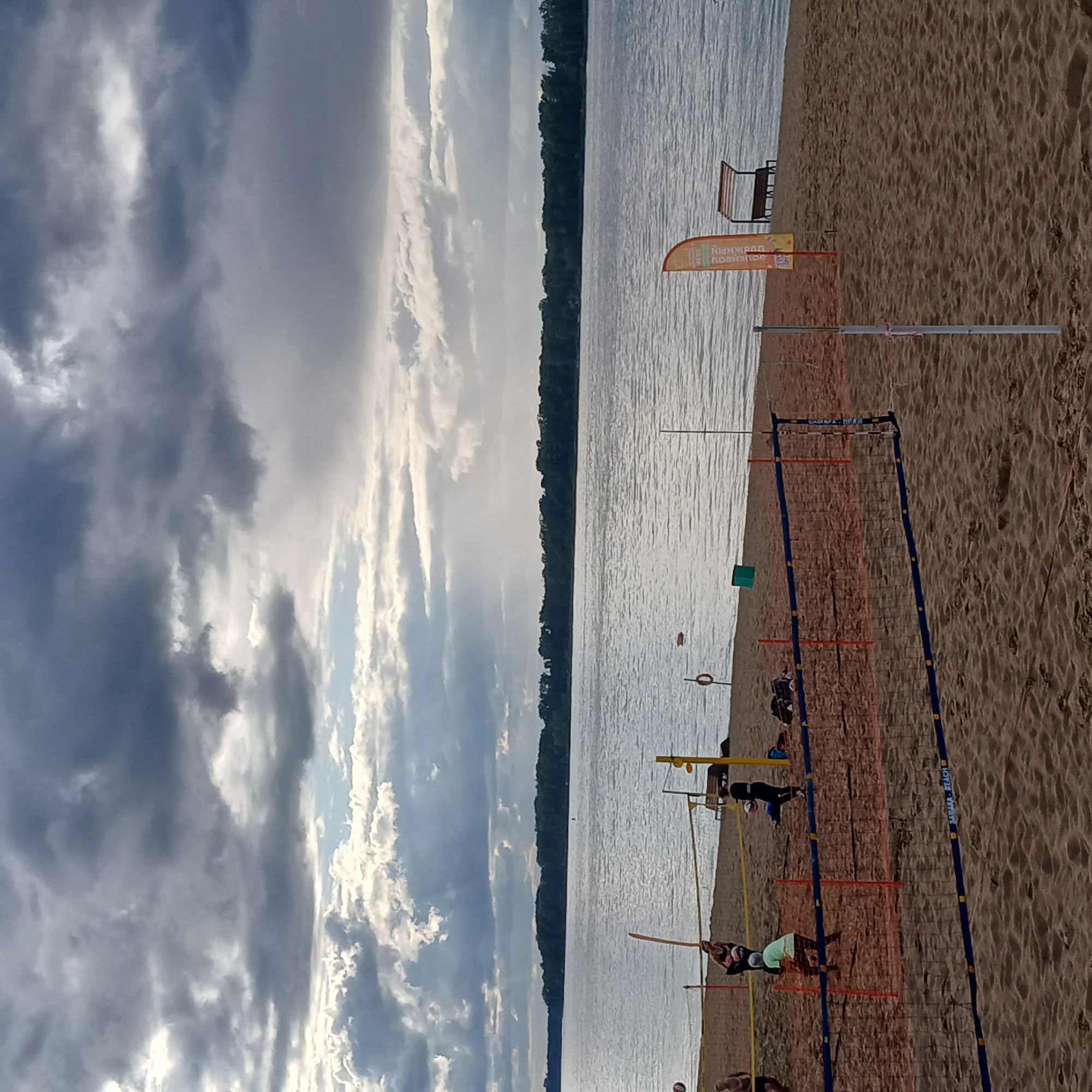 The image depicts a beach volleyball scene on a sandy shore. The sky is overcast with dramatic, dark clouds interspersed with patches of lighter sky, suggesting a change in weather. A wide body of water, likely the Volga River, stretches across the background, with a tree-lined shore on the opposite bank.
In the foreground, a beach volleyball net divides the sandy court. Several individuals are engaged in playing volleyball. On the left side of the net, a person in a green shirt is in motion, possibly preparing to hit the ball. To their right, another person in dark clothing is also actively participating in the game. On the far left, two other individuals are partially visible near the water's edge.
Various objects are present on the beach, including volleyball poles with nets, a small orange safety buoy in the water, a green trash bin, and a bench with a canopy structure on a slight incline. An orange flag with Cyrillic text is planted in the sand near the canopy. The sand itself shows footprints and the texture of a typical beach environment. The scene appears to be set in Samara, Russia, potentially at one of its designated beaches along the Volga River.