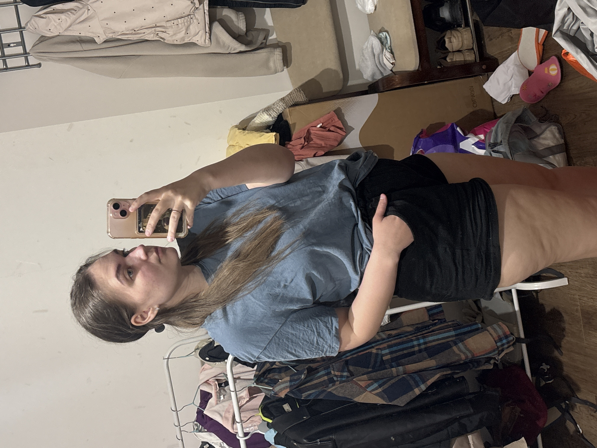 A woman is taking a selfie in a room with a messy closet. She is holding a phone in her right hand, angled to capture her face and upper body. She has light brown hair and is wearing a blue t-shirt and black shorts. Behind her is a clothing rack filled with various garments, including shirts and jackets. The room is cluttered, with clothes and other items strewn about on the floor and surrounding furniture. The location is not specified in the image.