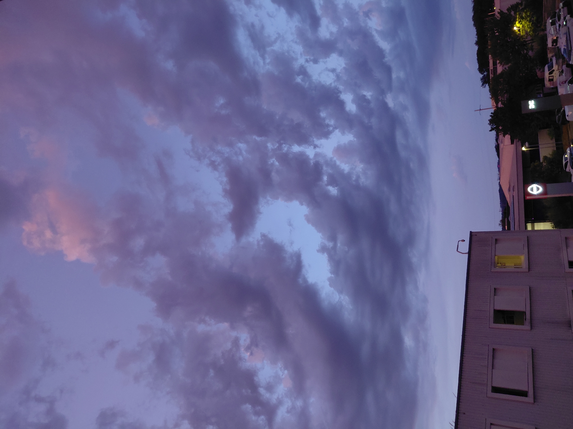 The image shows a view of Ecublens, Switzerland, during dusk. The sky is filled with dramatic purple and pink clouds, with a hint of lighter blue peeking through. In the foreground, a modern, light-colored, multi-story building with several rectangular windows is visible on the left. One window on the right side of this building is illuminated with a warm yellow light. In the background, a treeline and other buildings are faintly visible. A street with several parked vehicles, including vans and cars, is also present in the lower right portion of the image. A sign with a prominent 'e' logo is visible on the left side of the background, potentially indicating a commercial area.