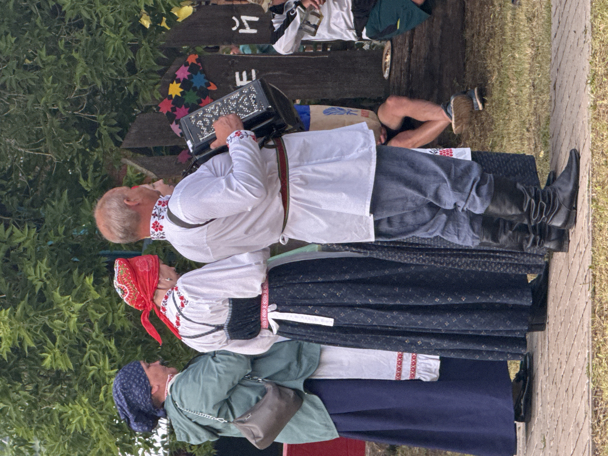 In Старые Быги, Russia, a group of people are gathered outdoors, possibly for a cultural festival or performance. The scene is dominated by three individuals dressed in traditional folk attire.
The person in the foreground, a man, plays an accordion. He wears a white shirt with embroidered red and black detailing around the collar, dark pants, and black boots. He is focused on his music.
Behind him, a woman in a white blouse with red embroidery and a dark, patterned skirt is visible. She wears a red headscarf with a vibrant pattern.
Further back, another woman is seen wearing a green jacket over a white blouse with red embroidery and a dark blue headscarf. A small, dark brown purse hangs from her shoulder.
The backdrop includes lush green foliage, a wooden fence with decorative elements, and a paved walkway. The overall atmosphere appears festive and communal. The time of day seems to be daytime, and the weather appears to be pleasant, suitable for an outdoor gathering.
Visible text includes stylized letters "Ч" and "Е" on wooden planks in the background, likely part of a sign or decoration. The overall impression is one of cultural celebration and musical performance in a traditional setting.