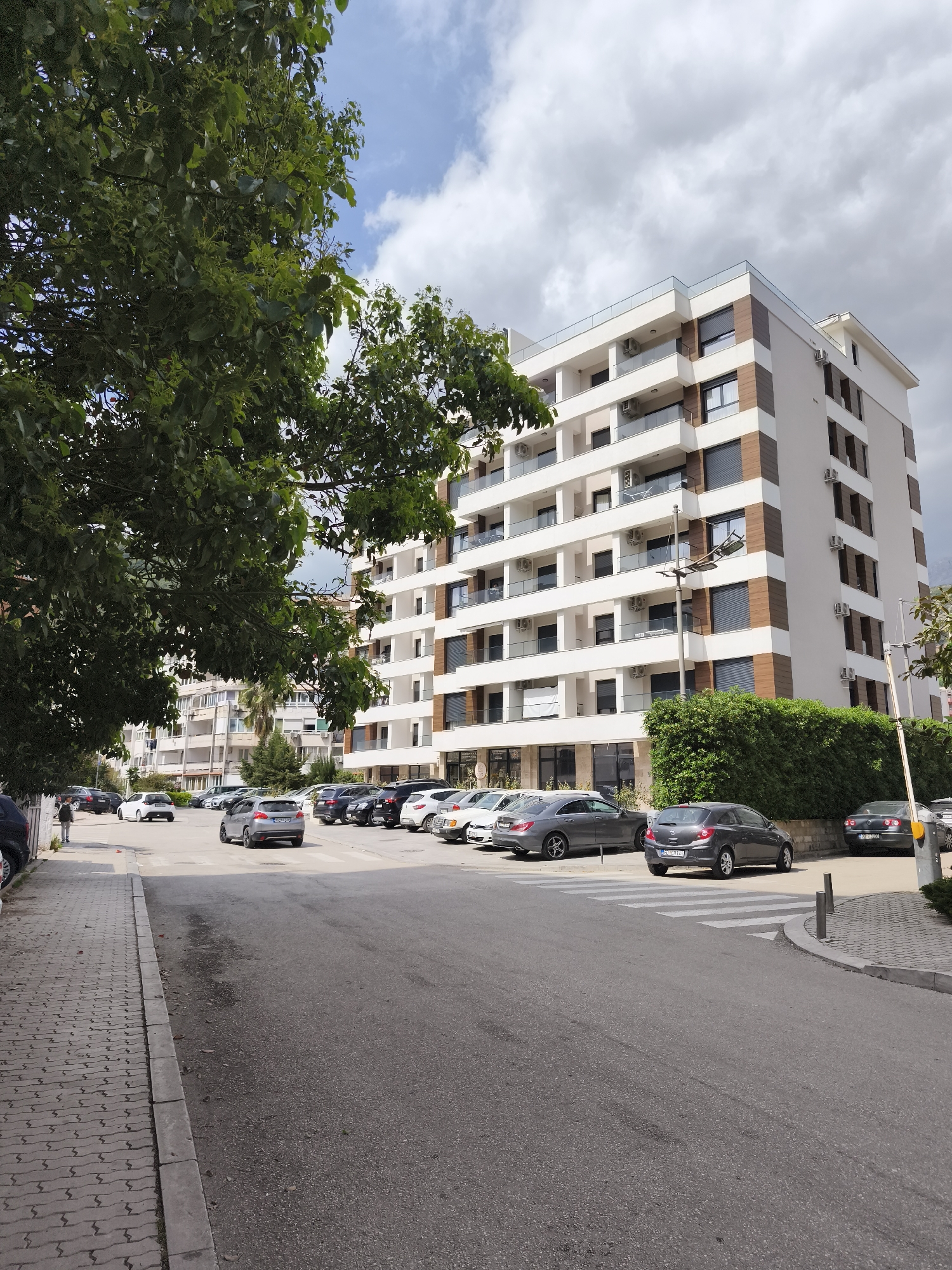 This image captures a bright daytime street scene in Budva, Montenegro, under a sky filled with scattered white and grey clouds. The main subject is a modern, multi-story building, likely residential, dominating the background. Its facade is a mix of white plaster and brown wooden-like panels, featuring numerous balconies with glass railings and individual air conditioning units. A green hedge runs along the base of the building, separating it from a parking area.
In the mid-ground, an asphalt road leads to a parking lot filled with various cars. A crosswalk is visible on the right side of the road. A dark grey Opel Corsa hatchback, bearing the Montenegrin license plate "HN CG 194", is prominent in the parking area. To the far right, another dark car with the license plate "BD 066" (indicating Budva) is partially visible behind a barrier. Several other cars are parked throughout the area.
On the left side of the frame, a brick-patterned sidewalk runs alongside a large, leafy green tree that partially obscures the view of the background. A single person is seen walking away from the camera on this sidewalk. A street lamp stands near the main building. The overall impression is an urban environment, likely a residential or mixed-use area, experiencing a typical day. A small, unreadable sign is visible on the facade of the main building.
