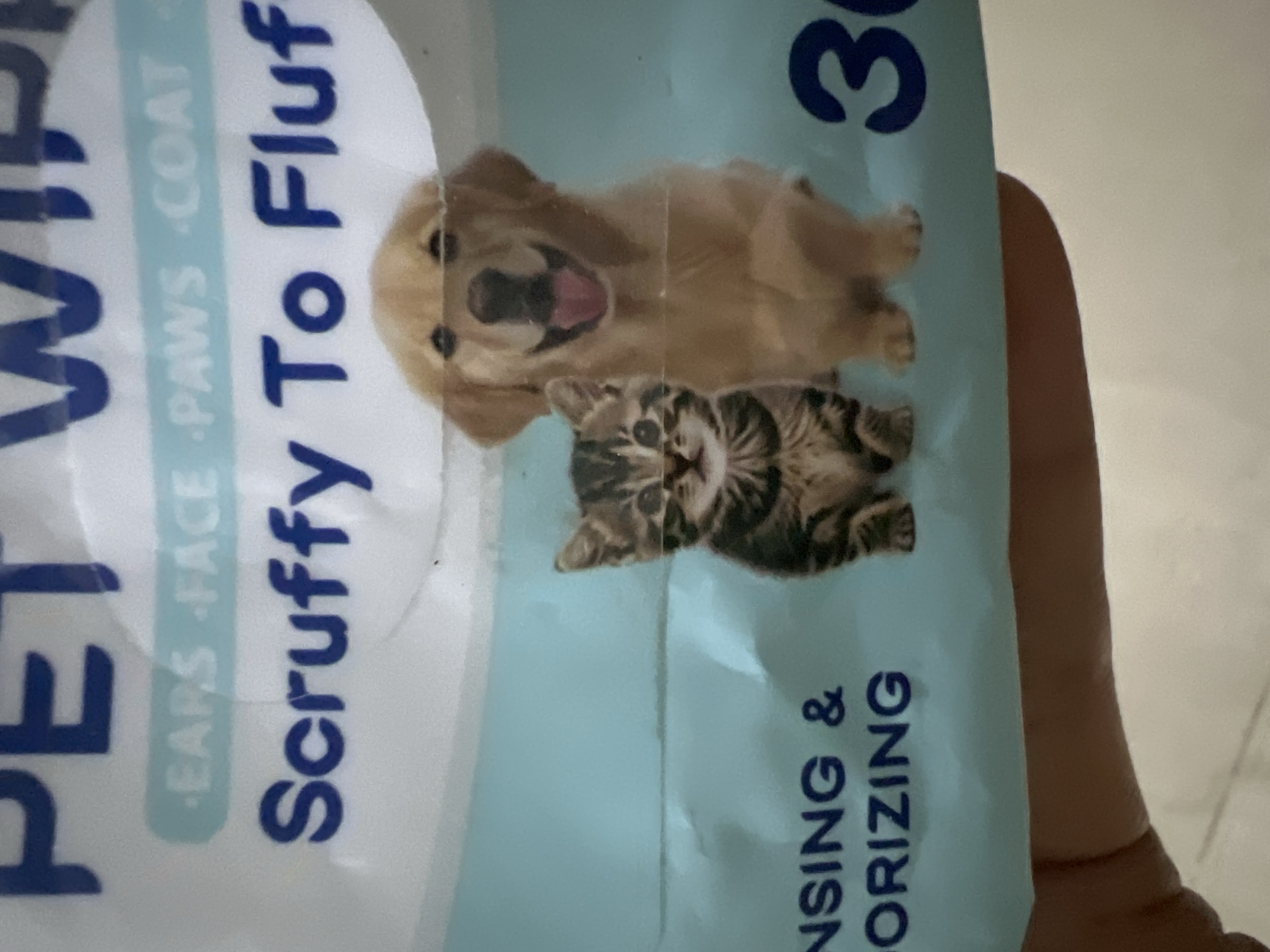The image shows a close-up of a package of pet wipes. The packaging features large blue text that reads "PET WIPES" at the top. Below this, in a lighter blue background strip, are the words "EARS FACE PAWS COAT". Centered on the package are images of a kitten and a golden retriever puppy. The kitten is on the left, looking forward with a striped tabby pattern. The puppy is on the right, with its mouth open in a happy expression, tongue visible. Below the animals, more blue text is visible, reading "NSING &". To the far right, a large blue number "30" is present. The background is a light blue. The image is taken from a low angle, with a hand partially visible in the lower left corner, holding the packaging. There is no identifiable location in the city of Klapanunggal, Indonesia.