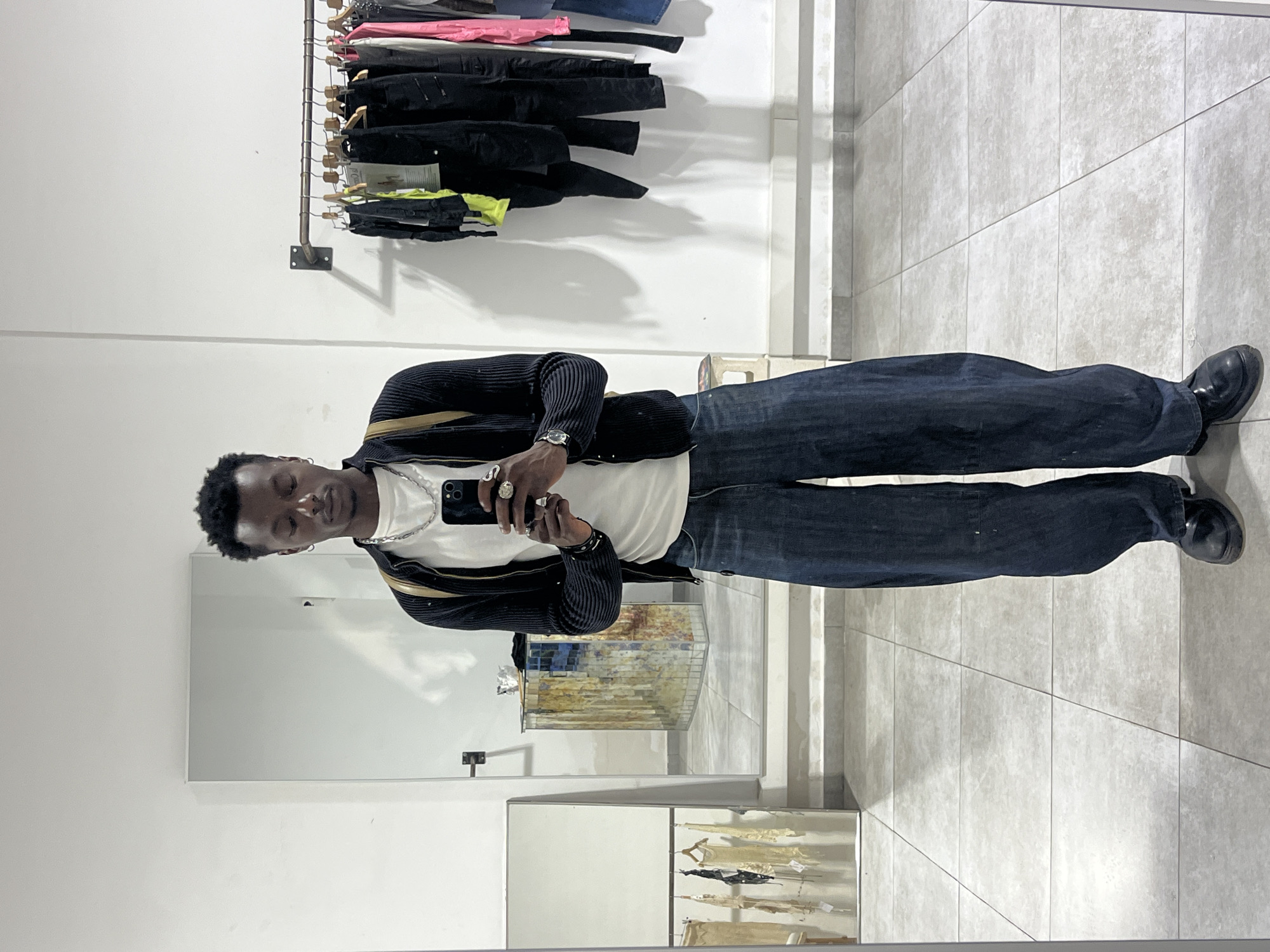 The image is a full-body self-portrait of a man taken in what appears to be a clothing store or boutique. He is facing the camera, holding a smartphone, likely to take the picture, and is standing in front of a large mirror that shows the reflection of the surrounding store. The man is wearing a white t-shirt, a dark cardigan with gold trim, dark blue jeans, and black shoes. He has a silver chain necklace, rings on his fingers, and a watch on his wrist.
Behind the man, clothes are displayed on a rack against a white wall. To the side, more clothes are seen on display in the reflection of the mirror. The floor is tiled in light grey. The environment suggests a modern, minimalist interior design.
Based on the presence of Catalan language signage in the background's reflection, it is likely that the location is in Barcelona, Spain. The man is not interacting with anyone else; he is solely focused on taking the self-portrait.