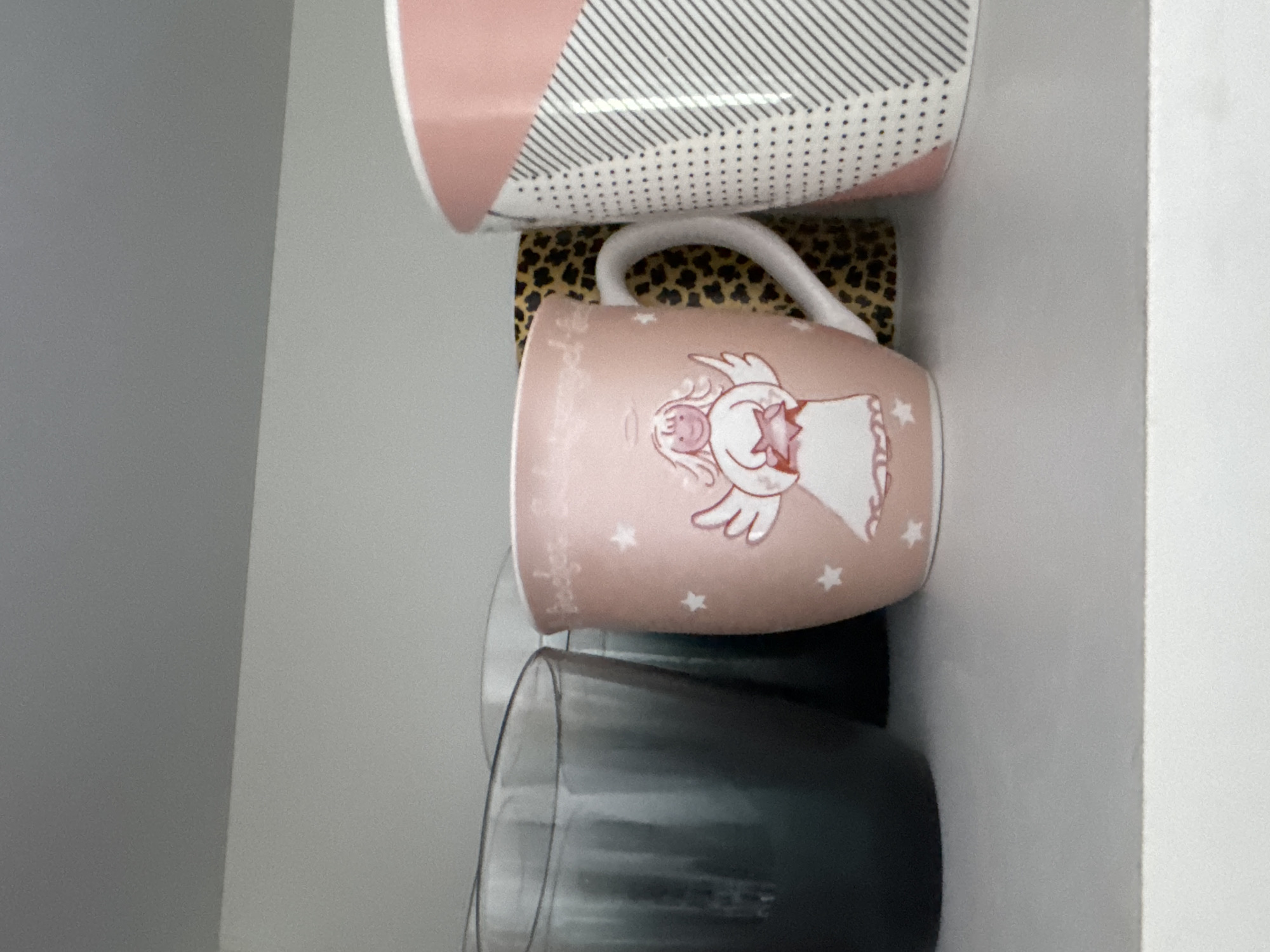 The image shows a shelf with several mugs and glasses. In the center is a pink mug with an illustration of an angel holding a star. To the left of the pink mug are two dark-colored glasses. To the right of the pink mug is a mug with a leopard print pattern, and further to the right is another mug with a geometric pink and white pattern. The shelf appears to be located inside a cabinet or cupboard, possibly in a kitchen or living area. The background is a plain, light-colored wall. No people are visible in the image. The scene is static and does not depict any action or interaction. The image quality is clear, and the objects are well-lit. This scene is not specific to any location in the city of Höhenkirchen-Siegertsbrunn, Germany, as it depicts a generic interior setting.