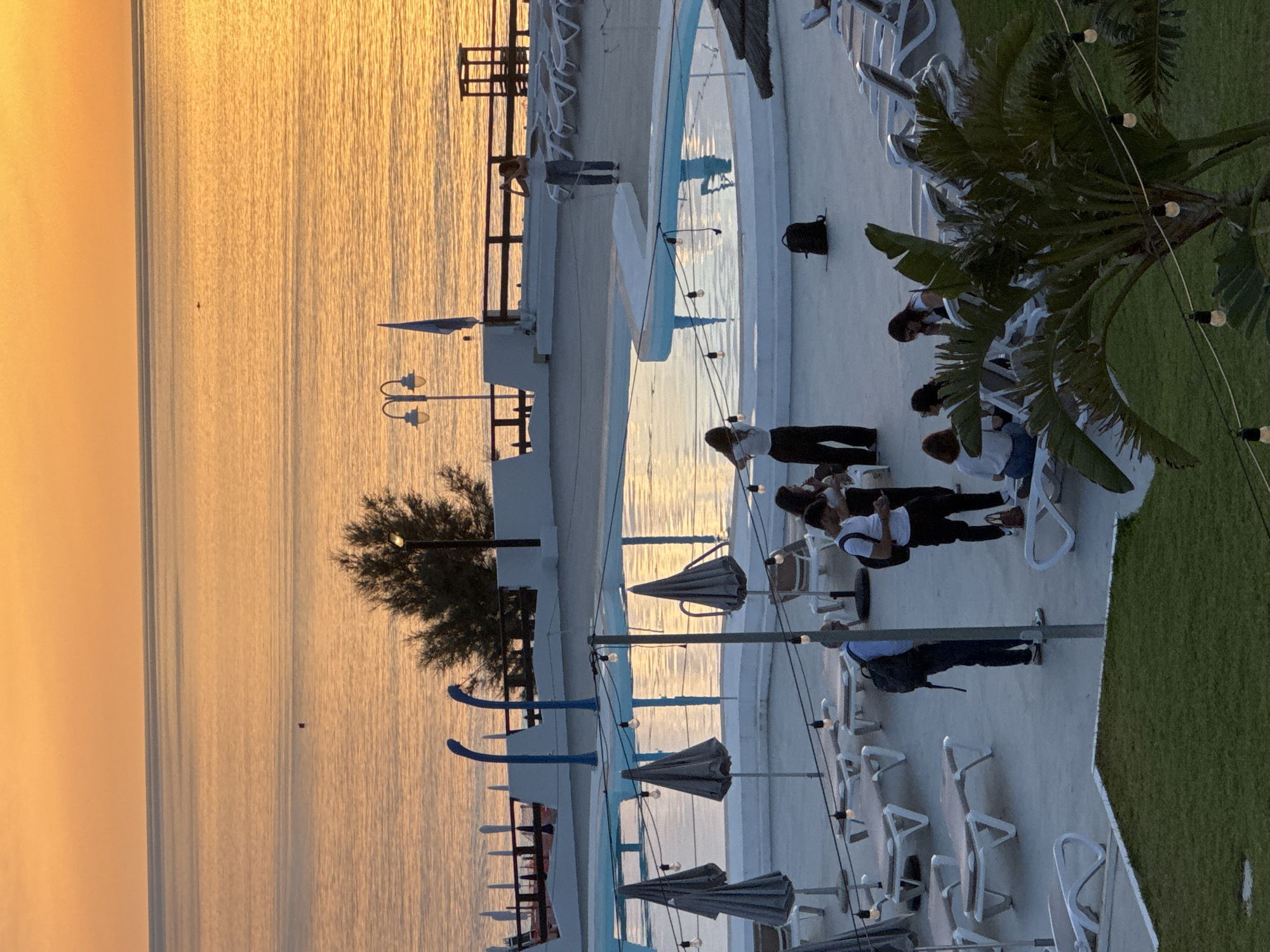 The image depicts a waterfront resort or pool area at sunset in Isola delle Femmine, Italy. The sky is a warm orange and yellow, reflecting on the calm sea. In the foreground, a white tiled deck surrounds a swimming pool, which is partially visible and reflects the sunset. Several lounge chairs and umbrellas are scattered around the deck, some occupied by people. A group of individuals are standing and sitting near the pool, some engaged in conversation, while others appear to be relaxing. In the background, a white railing separates the deck from the sea. A few palm trees are visible, adding to the tropical ambiance. A single red buoy is visible in the water.