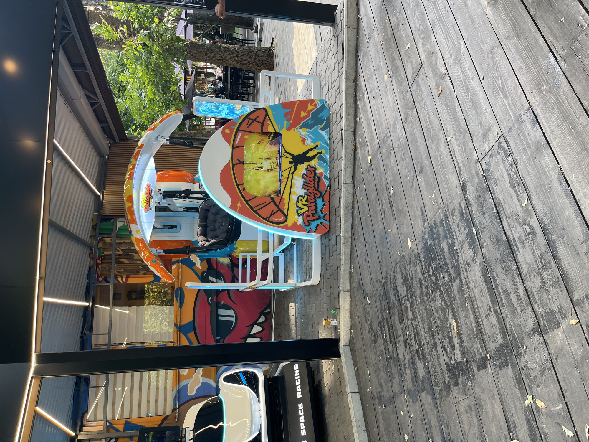 The image shows an outdoor amusement area, likely in Kharkiv, Ukraine, under a covered structure. In the foreground, a wooden deck with some debris and fallen leaves is visible. To the left, a white "VR SPACE RACING" simulator is partially visible. The central subject is a "VR Paraglider" attraction with a colorful, cartoonish design. It features a screen displaying a paragliding scene and a seated area with a harness. Behind the attractions, there is a wall adorned with graffiti depicting a red monster with sunglasses. Further in the background, trees, outdoor seating with umbrellas, and people can be seen, suggesting a public space like a park or a café. The lighting indicates it is daytime.