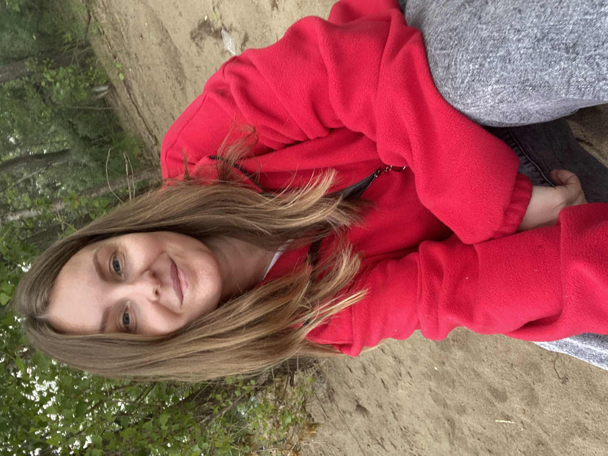 The scene is set outdoors in a natural environment, specifically identified as being in the city of Chaikovsky, Russia. A female individual, appearing to be an adult between 25 and 35 years of age, is positioned centrally, facing the camera directly. She has long, straight, light brown hair parted on the left, extending over her shoulders. Her eyes are blue/grey, and she displays a slight, closed-lip smile. She is wearing a bright red fleece hooded jacket, and grey pants or jeans are visible from the waist down. A black strap, possibly from a bag or camera, is discernible over her right shoulder, partially obscured by her hair and jacket. A metallic zipper pull or chain is present near the collar of her jacket. The individual is in a crouched or seated posture on light brown sandy or dirt ground, which covers the foreground and midground. The background consists of dense green deciduous foliage and multiple tree trunks, indicating a wooded or forested area. The lighting is natural and appears diffused, suggesting an overcast condition or a shaded area.