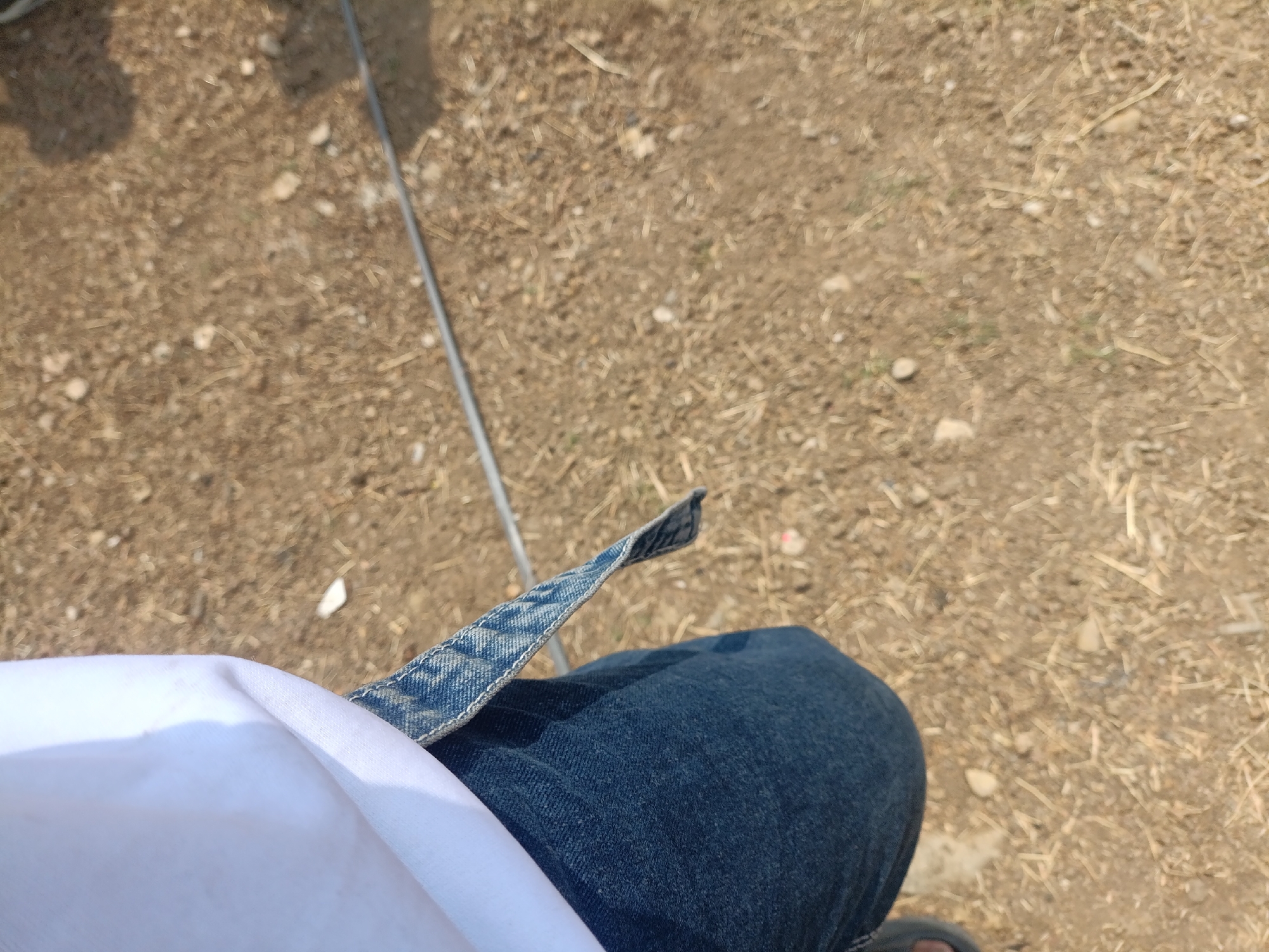 The image shows a person's lap and legs, wearing dark blue denim jeans and a white garment. A part of the denim jeans, specifically a pocket or belt loop area, is visible with visible stitching and texture. The background consists of dry, brownish ground with small stones and scattered dry vegetation, indicative of an outdoor environment. There is a thin, metallic or plastic rod extending diagonally from the upper left to the middle of the frame, casting a shadow. The overall lighting suggests it is daytime. There is no visible indication of the specific location within Cariu, Indonesia.
