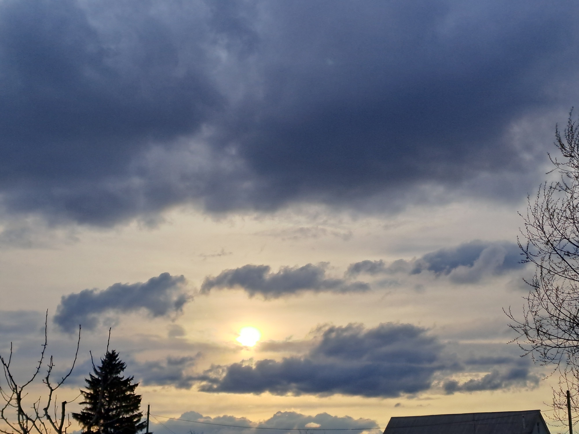 The image displays a serene yet dramatic sky scene, likely captured during sunrise or sunset, over Polyanki, Russia. The upper half of the frame is dominated by heavy, dark grey and blue clouds, creating a sense of depth and impending weather. Below these denser formations, the sky transitions into lighter shades of pale yellow, orange, and light grey.
A bright, intense yellow sun is visible in the middle-lower section of the sky, partially obscured by scattered, elongated grey clouds. The warm glow of the sun contrasts sharply with the cooler tones of the clouds.
Along the bottom edge of the frame, silhouettes of trees and a building are visible. On the far left, bare, branching limbs of a deciduous tree reach into the sky. Below this, a dark, prominent coniferous tree, likely a pine or spruce, stands out. More bare tree branches are present on the right side of the image. The roofline of a simple building, possibly a house or barn with a dark or grey roof, is visible in the bottom right corner.
The bare branches suggest the season is late autumn, winter, or early spring. The interplay of strong sunlight with varied cloud formations creates a captivating and atmospheric landscape. There is no visible text in the image.