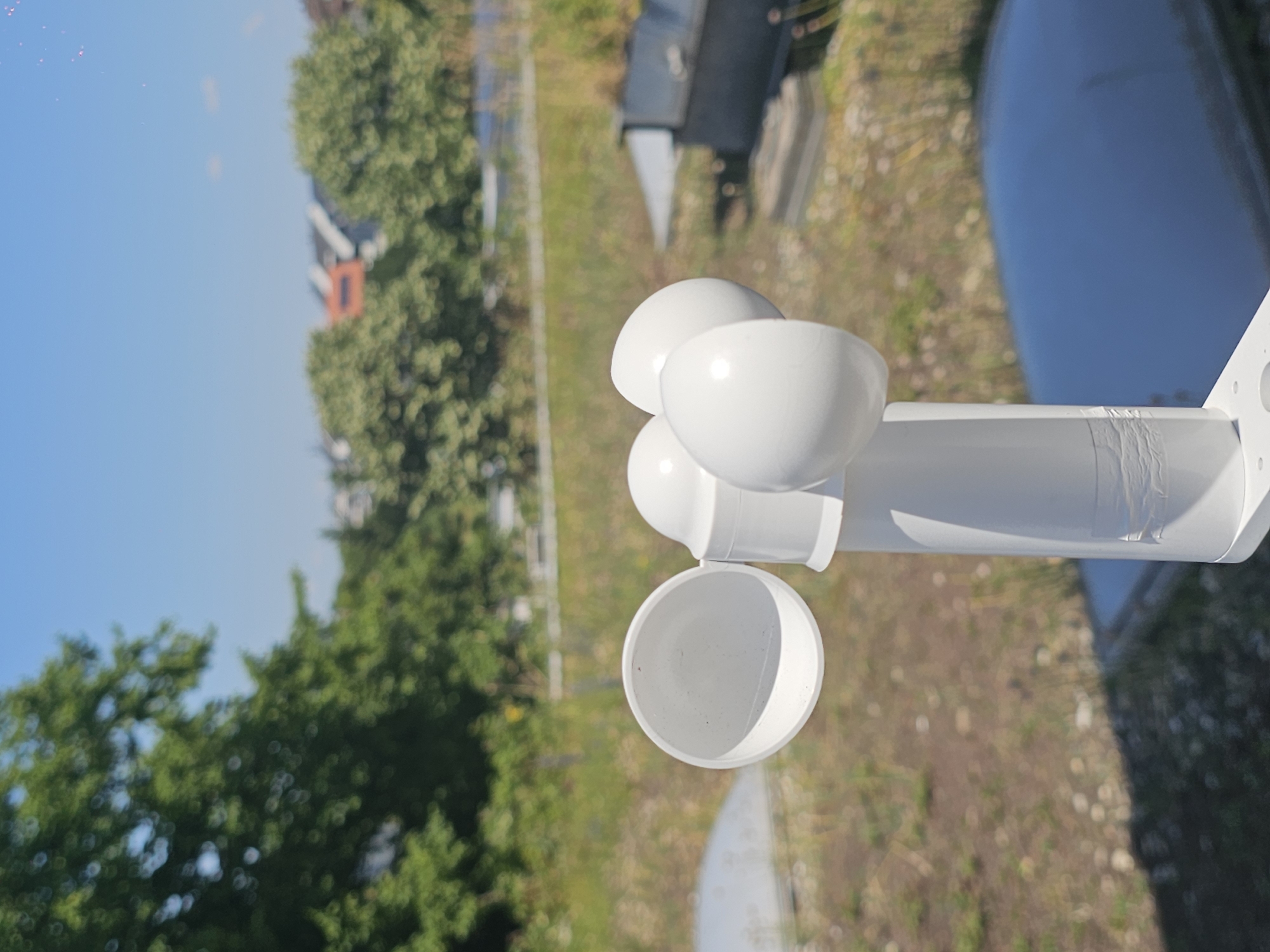 This is a media file depicting a close-up view of white hemispherical cups attached to a white cylindrical object. The cups appear to be part of a weather instrument, possibly an anemometer or a rain gauge. The image is shot from a slightly downward angle, with the cups prominently featured in the foreground.
The background is out of focus but suggests an outdoor setting. Lush green trees are visible on the left side, and a light blue sky occupies the upper portion of the image. The ground in the background appears to be covered with dry grass and small white flowers. A body of water, possibly a pond or a lake, is reflected in the shiny surface to the right of the cups.
The lighting suggests it is daytime, with bright sunlight illuminating the scene, creating highlights on the white cups. The overall mood is bright and clear, indicative of fair weather. There are no people visible in the media file, and no text is present. The context of the location is unknown, but the presence of trees, grass, and water suggests a natural or semi-natural environment. The focus on the weather instrument hints at an interest in meteorological data or observation.