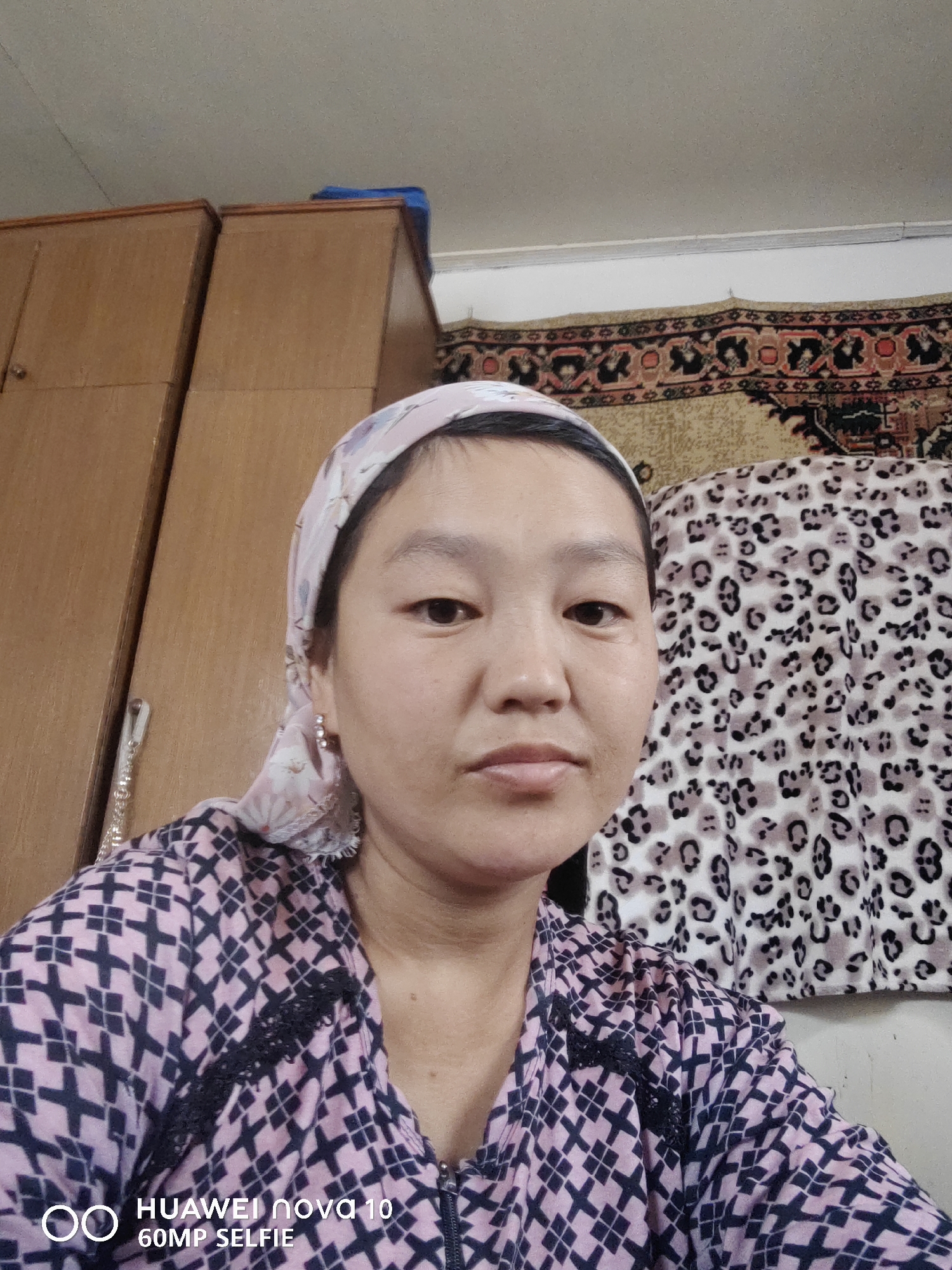A close-up, head-and-shoulders selfie of a person wearing a pink and navy patterned shirt and a pink headscarf is presented. The person has dark hair, brown eyes, and is looking directly at the camera with a neutral expression. To the left of the person, two brown wooden closet doors are visible, and a silver chain hangs from one of them. The background includes a tapestry with a red and black geometric pattern, and a leopard-print fabric. The text "HUAWEI nova 10 60MP SELFIE" is overlaid in the lower-left corner of the image. The scene appears to be indoors, possibly in a domestic setting in Jetim-Dobo, Kyrgyzstan.