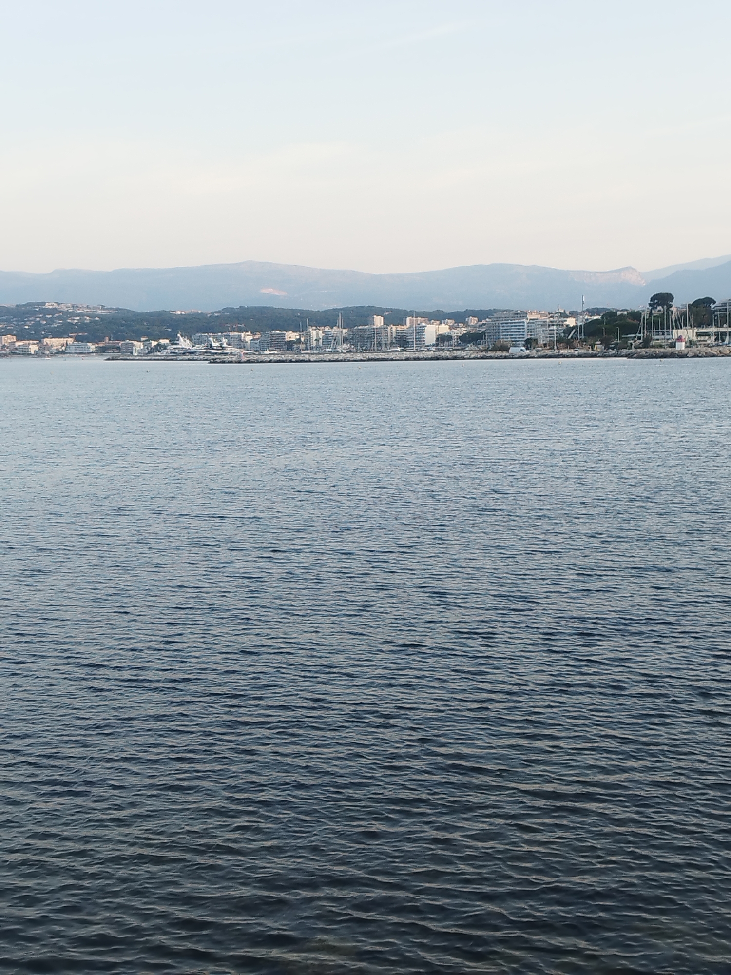 This is a high-angle, full shot of a body of water in front of the city of Antibes, France. The water is dark blue and choppy, with small waves. In the background, the city of Antibes is visible, with buildings, trees, and a mountain range. The sky is light blue and clear. There are no people or animals visible in the image. The scene appears to be peaceful and serene.