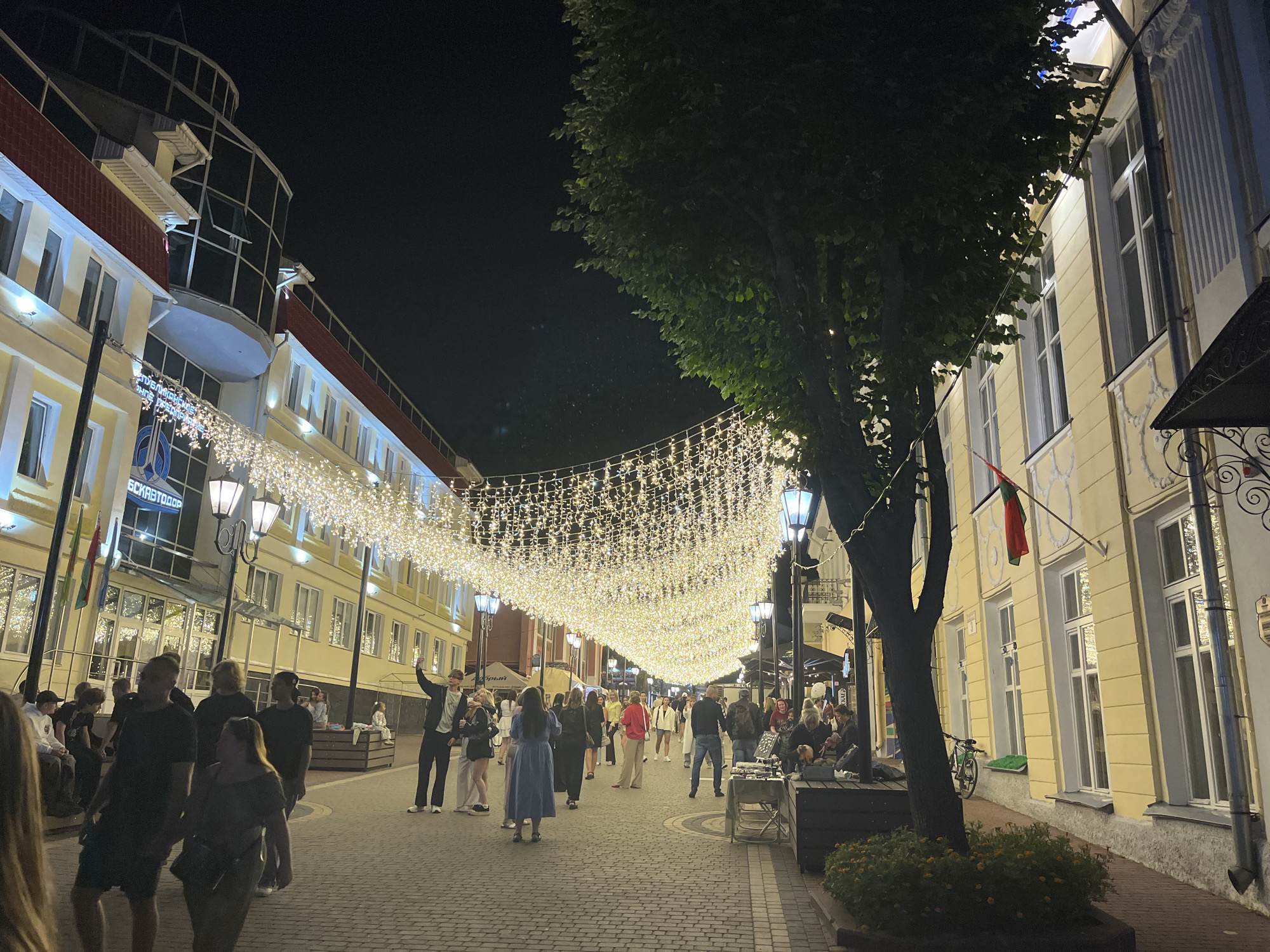 The image depicts a pedestrian street at night, illuminated by strings of warm white lights strung across its width. The street is lined with multi-story buildings. On the left, a building with a yellow facade, red roof accents, and a modern glass-enclosed upper section is visible. A sign on this building, partially obscured by lights, appears to be in Cyrillic script. On the right, a building with a yellow facade and numerous windows is shown, with a dark awning extending from its front. A large tree with dark green foliage is positioned in the foreground on the right, its branches partially obscuring the building.
The cobblestone street is populated by a diverse group of people. Several individuals are walking, some in pairs and others in small groups. Some are engaged in conversation, while others appear to be strolling or taking in the ambiance. A few people are seated on benches or low walls. In the foreground on the left, a person's arm and shoulder are visible, suggesting the photographer is among the crowd. A man in a dark suit and white shirt is raising his arm, holding a phone, potentially taking a photo or video. A bicycle is leaning against the wall of the building on the right. Several street lamps with decorative fixtures are also present along the street. The overall impression is a lively, festive evening atmosphere. The location is the pedestrian street on Suvorov Street in Vitsebsk, Belarus.