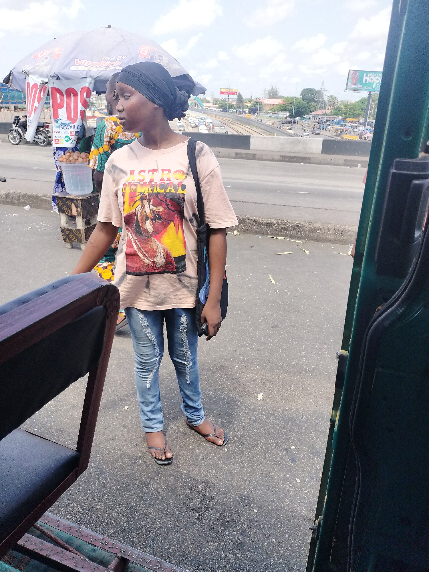 This street-level photograph captures a young woman standing next to a roadside vendor stall in a bustling urban setting, consistent with Lagos, Nigeria. The woman is wearing a black headwrap, a light-colored t-shirt with a "ASTRO" graphic featuring a woman's face, ripped blue jeans, and flip-flops. She carries a black shoulder bag and holds a mobile phone, looking contemplatively towards her right.
Behind her, a makeshift stall is shaded by a grey umbrella displaying text such as "Plus B9," "Virakich," and "MIL." A banner on the stall prominently advertises "POS IS AVAILABLE," alongside mentions of "Gotv" and "GLO." Products for sale include a clear bucket of what appears to be fried dough balls and another containing water, resting on wooden crates. A person wearing a patterned shirt is partially visible behind the stall.
The background reveals a busy multi-lane road or highway, complete with an overpass and various vehicles, including a parked motorbike. A billboard in the distance partially reads "RENEW Hoo." The photograph seems to have been taken from inside a dark green vehicle, with its frame and a seat visible in the foreground, suggesting a perspective from a tricycle or similar transport. The daytime scene is characterized by an overcast sky, casting soft, diffused light over the vibrant street activity.