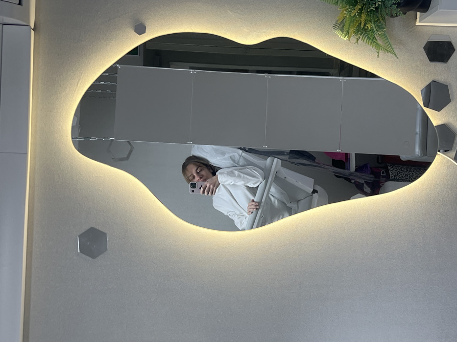 An image captures a female individual taking a mirror selfie in an indoor setting located in Ust-Ilimsk, Russia. The subject, with light brown hair, is seated in a white or light grey reclining chair, possibly of a medical or salon type, wearing a long-sleeved white garment. She holds a smartphone with a pink case in her right hand, positioned to photograph her reflection in a large, irregularly shaped mirror. The mirror features integrated backlighting, casting a warm glow on the surrounding textured, light grey wall. Several small, hexagonal, reflective decorative elements are adhered to the wall around the mirror.
The reflection within the mirror reveals a portion of the room behind the individual. To her left (viewer's right in the reflection), dark items, possibly bags or clothing, are visible on what appears to be a shelf or floor. To her right (viewer's left in the reflection), a large, light grey, multi-paneled partition or wall is reflected. Above this partition, multiple chain links are suspended from the ceiling. In the lower right quadrant of the mirror's reflection, a small potted plant with green foliage rests on a white surface. The overall environment suggests a modern clinic, spa, or beauty salon due to the specialized chair and contemporary decor.