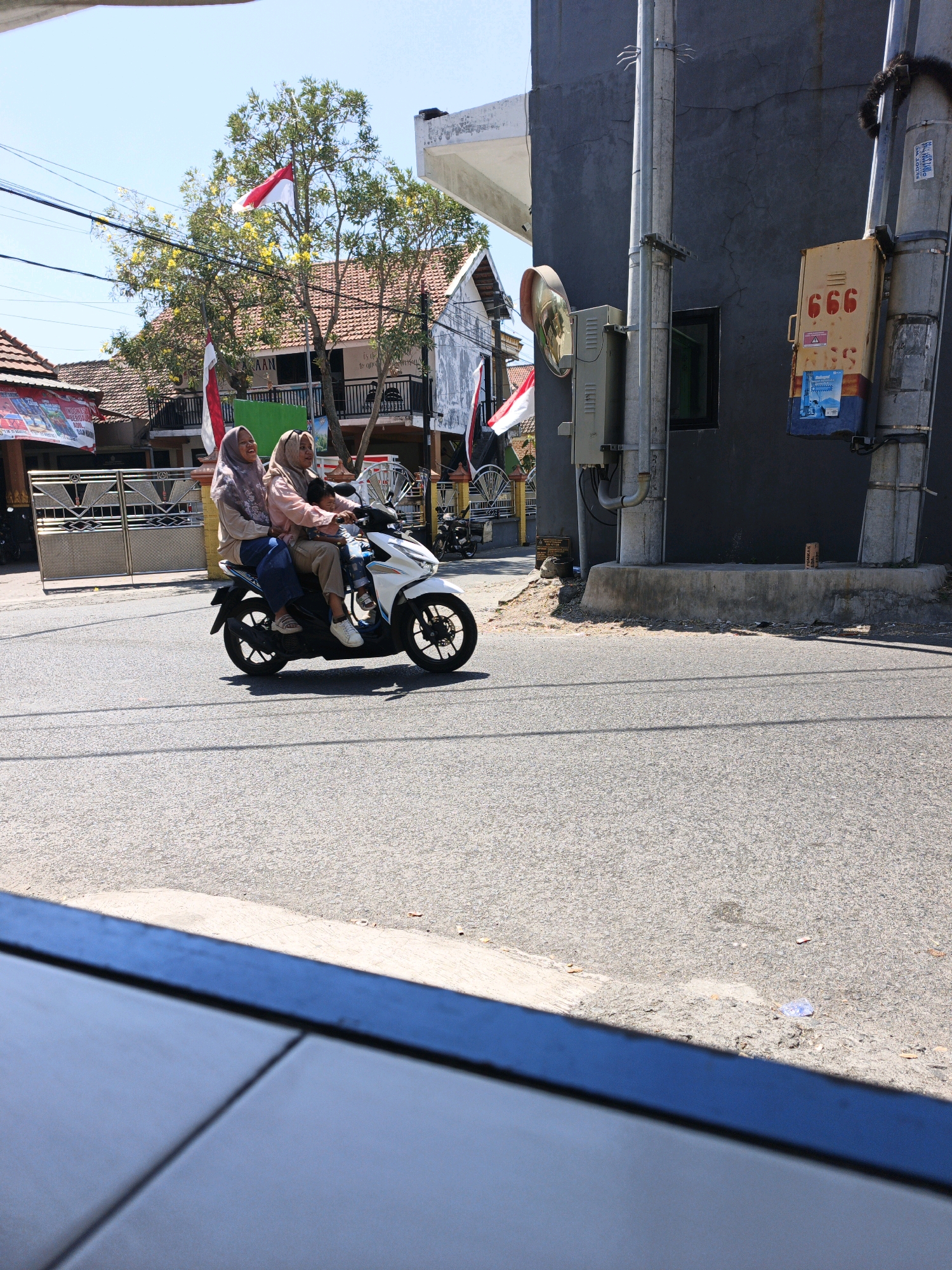 In Ketegan, Indonesia, two women and a young child are riding a white scooter on a paved road. The woman in the front is wearing a light pink hijab and a pink shirt, while the woman behind her wears a mauve hijab and a mauve shirt. The child, dressed in blue, sits between them. The scooter has black wheels and a white body. Behind the scooter, a grey building stands with a utility pole attached to its exterior, featuring a yellow box with the number "666" printed in red. In the background, trees with yellow flowers and buildings with red tile roofs are visible under a clear blue sky. Indonesian flags are displayed on poles. The road is asphalt, with visible tire marks and some scattered debris. In the foreground, a portion of a blue and grey surface, possibly a table or railing, is visible.