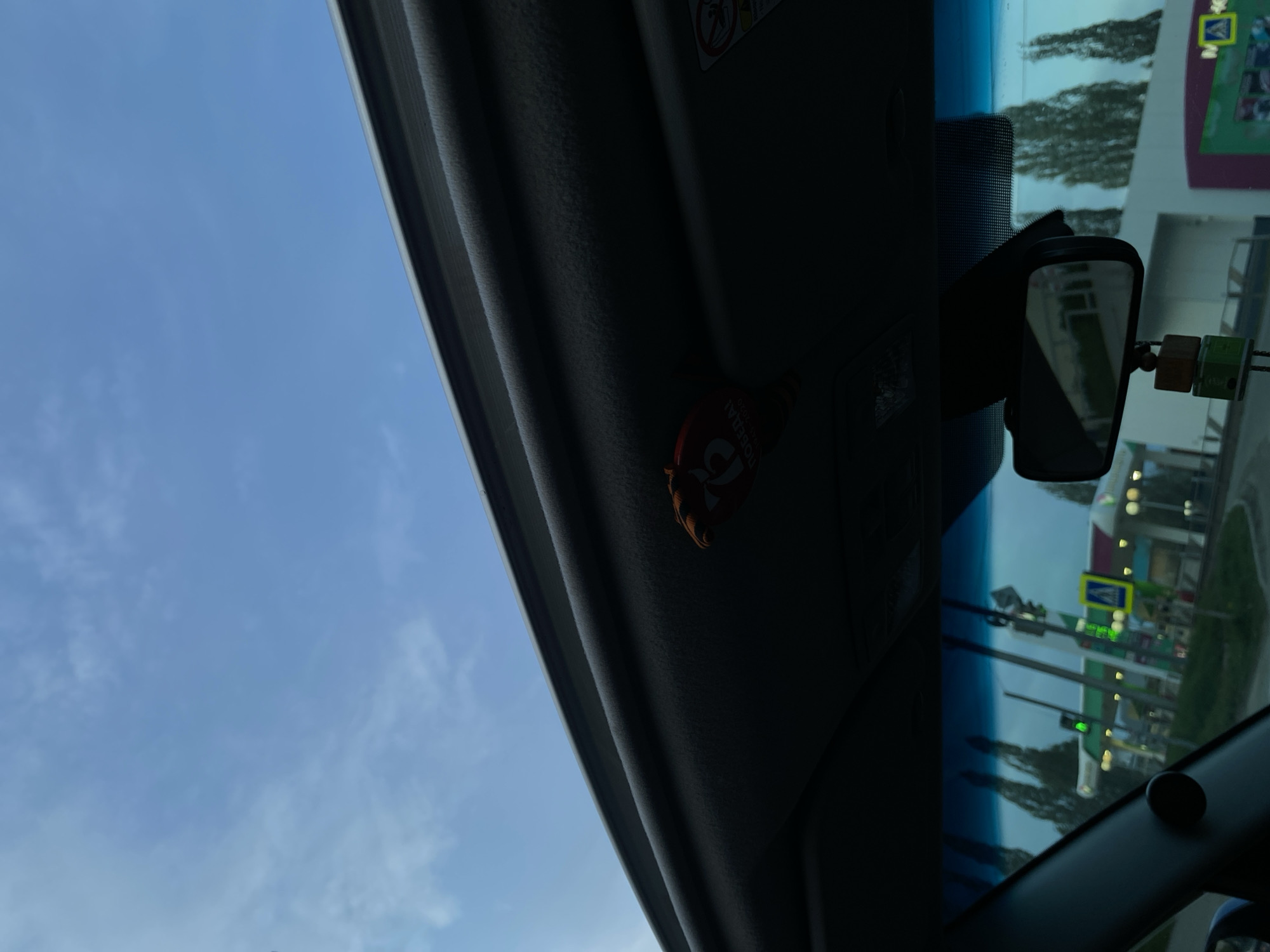 The image is taken from inside a vehicle, looking through the windshield. The sky is visible to the left, a light blue with some cloud cover. The dashboard and interior details are silhouetted in the foreground. A rear-view mirror and a small, likely decorative object are attached to the windshield. Through the windshield, the reflection shows an outdoor scene with trees, buildings, and traffic signs. The reflected scene is a streetscape and suggests the city of Votkinsk, Russia, based on the user's specification. There are no visible people, specific actions, or interactions in the image.