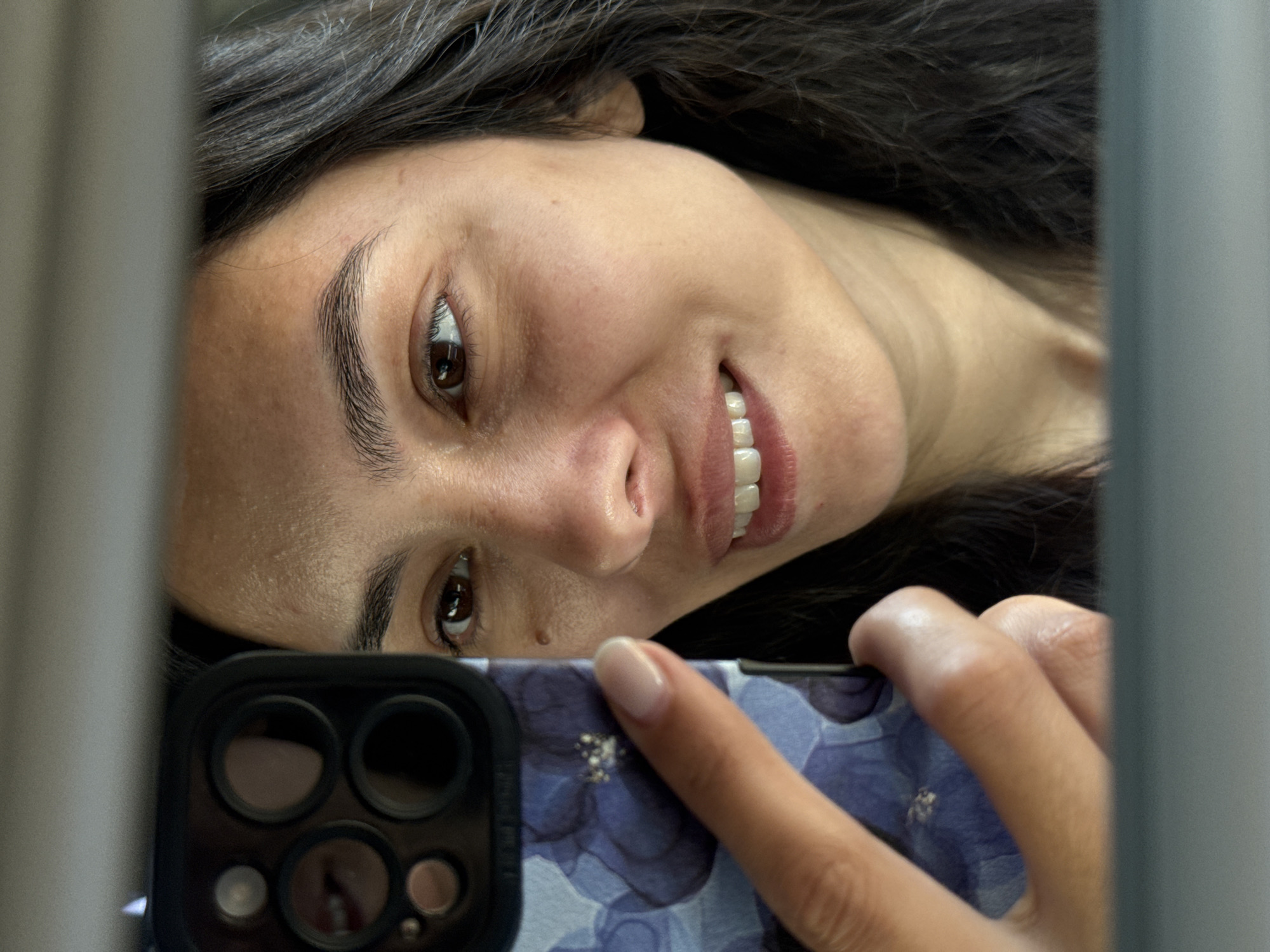 A woman is reflected in a mirror, holding a phone with a floral case in her left hand. She is looking at the phone, and a portion of her face is visible, including her right eye, nose, and mouth. The background is blurred. The image is a close-up shot and does not depict any specific location in the city of Chaikovsky, Russia.