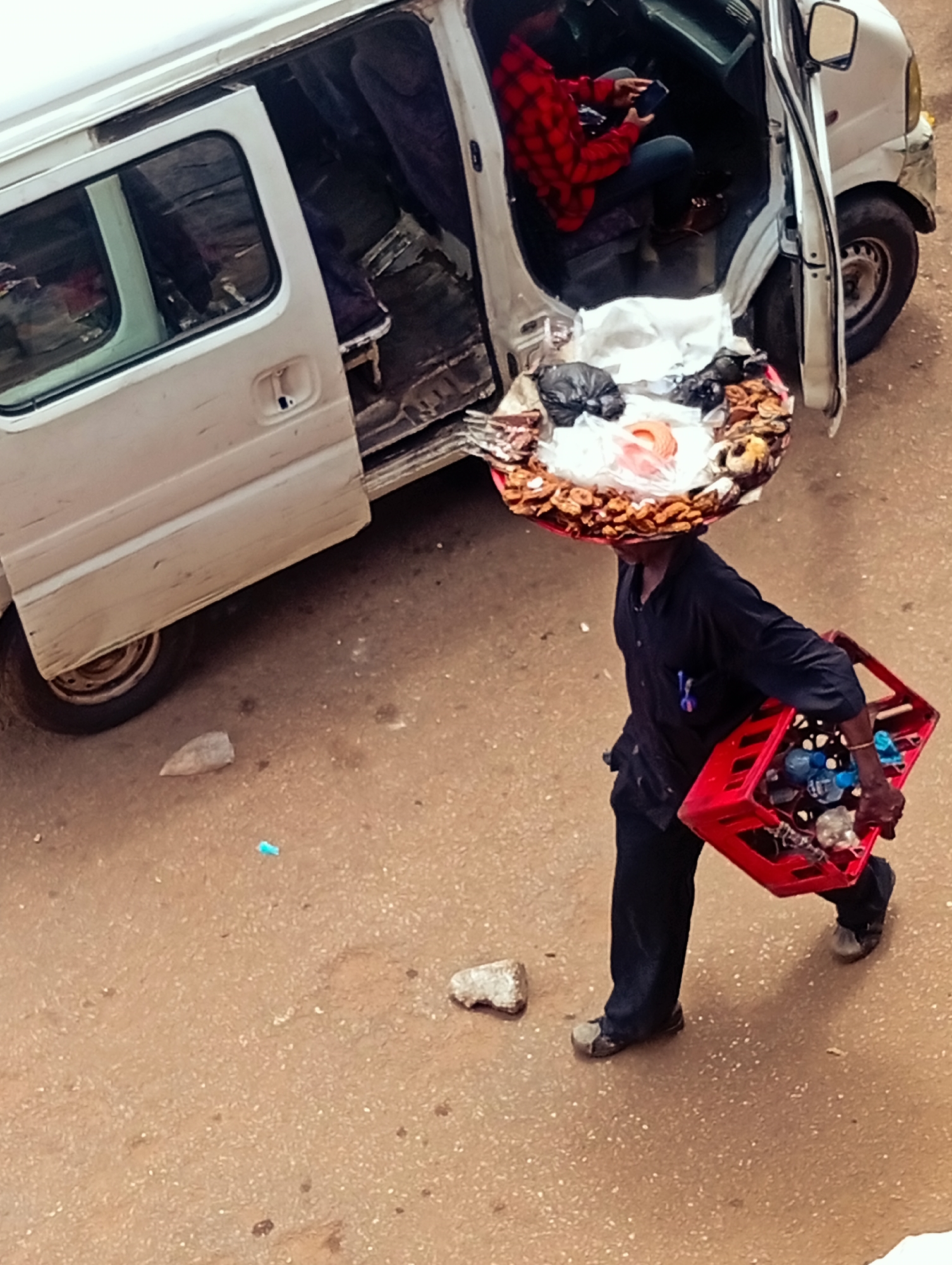 In Bukuru, Nigeria, a man walks on a dusty, paved surface carrying a red plastic crate filled with bottled drinks and other items. He is wearing dark clothing and carrying a large, round tray of food items balanced on his head, including what appears to be dried fruit and black plastic bags. To his left, a white van with its front passenger door open is parked. Inside the van, a person wearing a red and black checkered shirt is seated and looking at a dark-colored mobile phone. The van's interior is cluttered with various items.