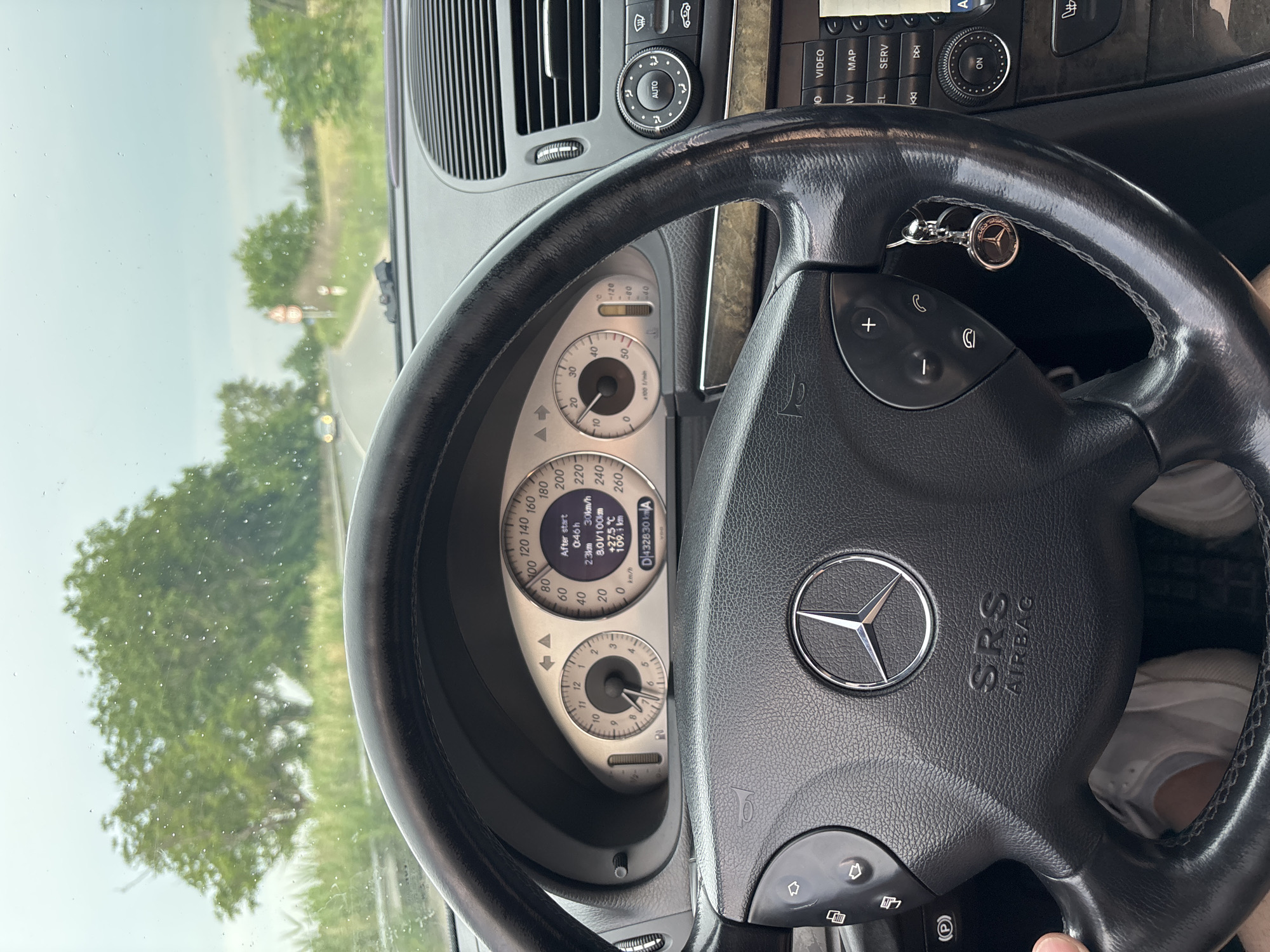 The image shows the interior of a Mercedes-Benz car, likely a W211 E-Class, from the driver's perspective. The steering wheel, dashboard, and instrument cluster are clearly visible. The instrument cluster displays information such as speed (30 km/h), engine temperature (+27.5°C), and odometer reading (432830 km). There is a key fob with a Mercedes-Benz logo hanging from the steering wheel. The car appears to be driving on a road with trees and vegetation on either side. A speed limit sign (60) is visible on the right side of the road. The sky is overcast, and there are raindrops on the windshield. Based on the Google Street View imagery of Yoakim Gruevo, Bulgaria, the road visible in the image is likely to be within the vicinity of the town, possibly on the road leading towards or away from it. The landscape suggests a rural or semi-rural setting. No people are visible in the image, other than the implied presence of the driver.