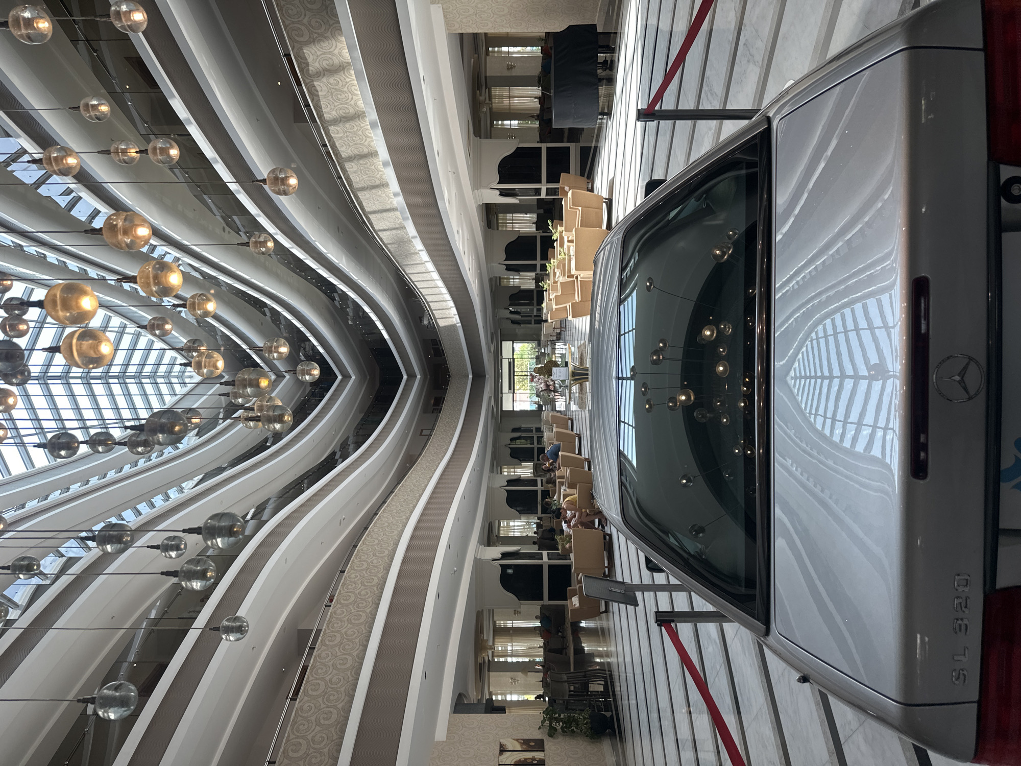 The image captures the interior of a large, multi-level atrium, likely a hotel lobby, in Manavgat, Turkey. The architecture features curved balconies and a glass ceiling that allows natural light to illuminate the space. Hanging glass pendant lights, some illuminated, are suspended at various heights, creating a dramatic effect. In the foreground, the rear of a silver Mercedes-Benz SL 320 is prominently displayed, partially obscuring the view of the lobby. The car's polished surface reflects the overhead lighting and the architectural elements of the atrium.
The lobby area is furnished with light-colored seating arrangements, including sofas and armchairs, arranged in conversation groupings. Several individuals are present in the background, engaged in activities such as sitting and conversing, suggesting a relaxed atmosphere. Decorative plants and floor lamps are visible, contributing to the overall ambiance. The floor is a polished marble with a striped pattern, and red velvet ropes on stanchions delineate areas of the space. In the far background, a grand piano covered with a black cloth is visible. The overall scene suggests a luxurious and well-maintained public space.